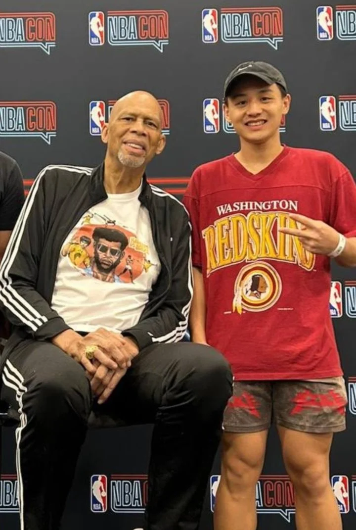 Two men posing at an NBA Con event, with a backdrop featuring NBA and NBA Con logos. The man on the left is bald, wearing a black Adidas jacket and black pants, sitting with his hands clasped. The man on the right is wearing a Washington Redskins t-shirt and camouflage shorts, standing and making a peace sign.
