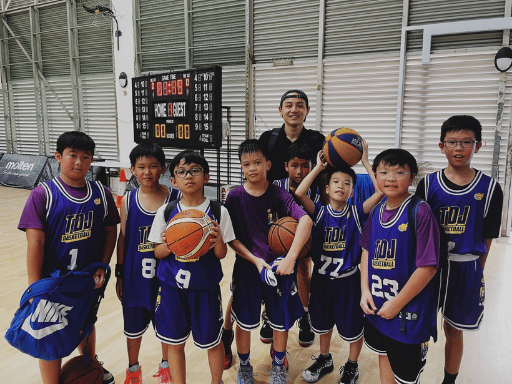 A group of young boys in purple and white basketball uniforms holding basketballs stands with a coach in an indoor basketball court. A scoreboard is visible in the background. TDJ U12 Team