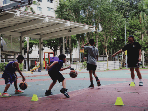 Children practicing basketball drills on an outdoor court, with cones set up for drills and a coach supervising.