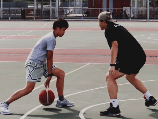 A young boy dribbling a basketball on an outdoor court while facing an older player in black athletic clothing, both in a defensive stance.