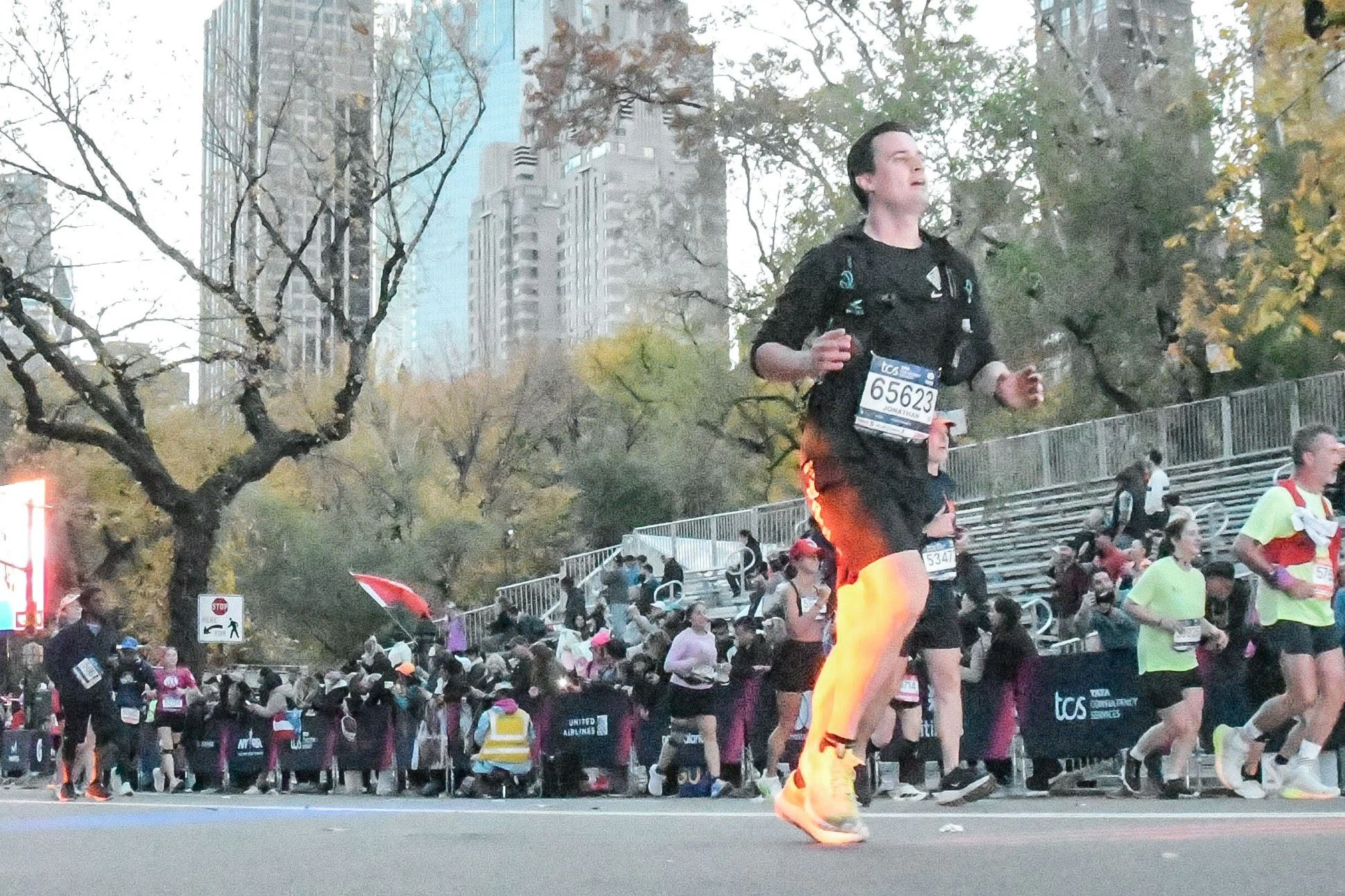 A group of runners participating in a marathon or race, running along a city street lined with spectators and trees with autumn foliage. In the background, there are tall buildings and a crowd of people watching the race.