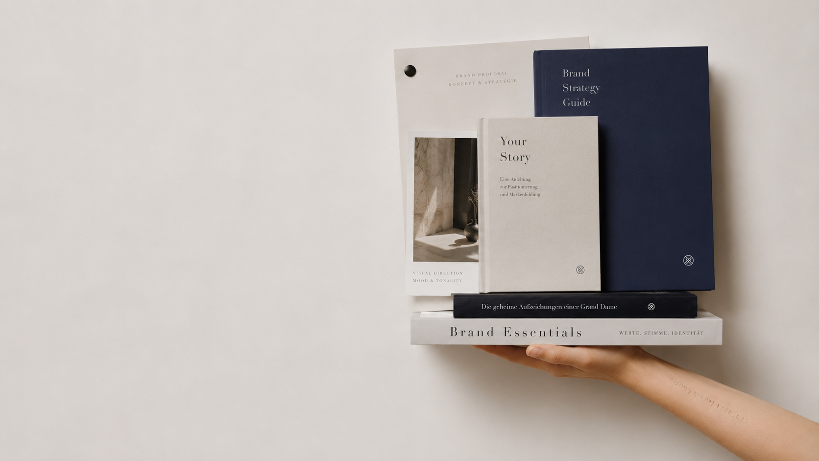 Eine Hand hält einen weißen Regal mit mehreren Büchern und Heften in neutralen Farben wie Weiß, Grau und Dunkelblau. Die Bücher und Hefte tragen Titel wie "Brand Strategy Guide" und "Your Story".