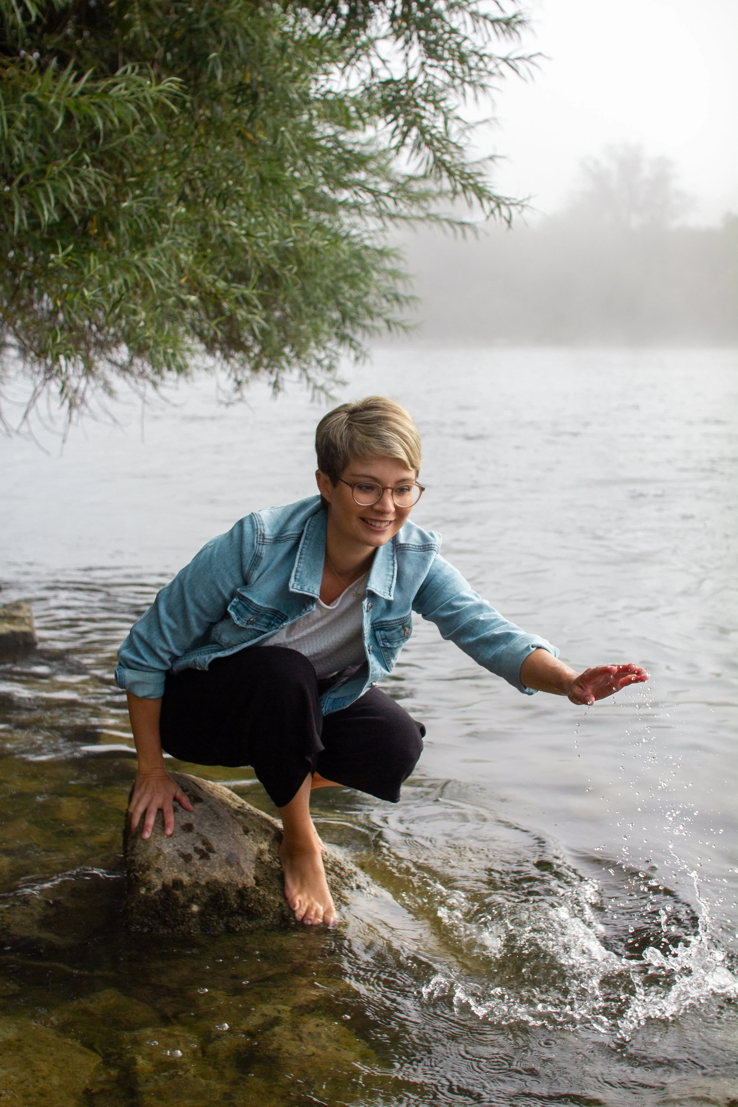 Junge Frau mit Brille, in Jeansjacke und schwarzen Hosen, kniet im Fluss und berührt das Wasser, umgeben von Nebel und grünen Bäumen.