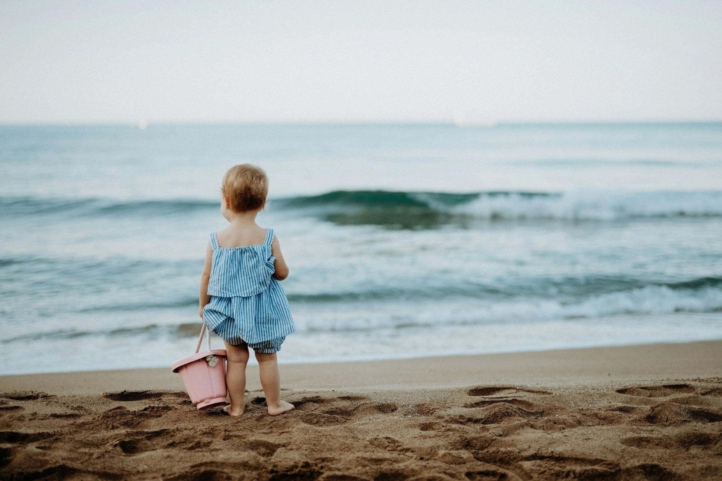 Kleines Mädchen mit blauer Strandkleidung steht am Strand, blickt auf das Meer, hält einen kleinen Korb.