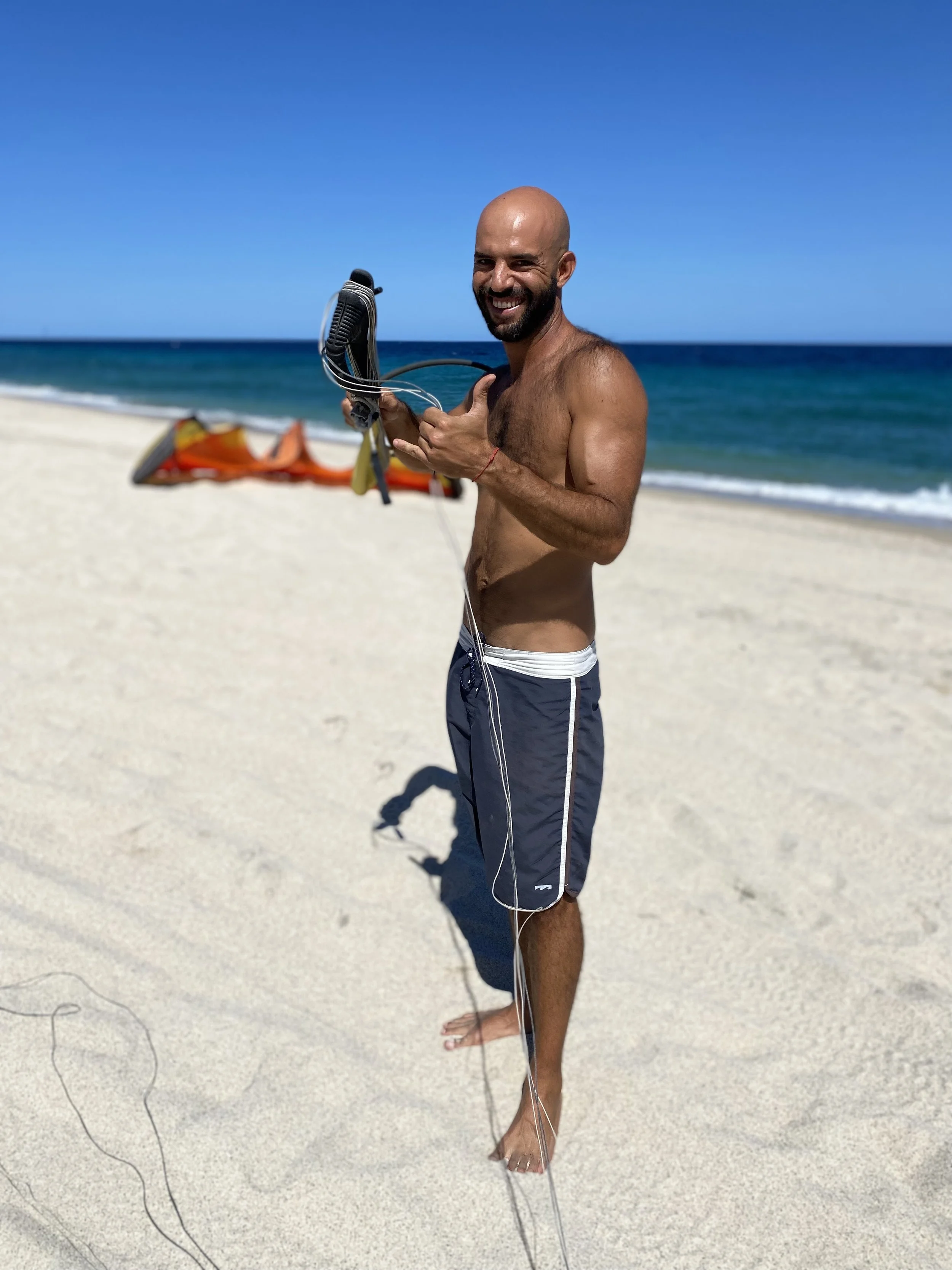 A person in board shorts on a beach, holding a kite control bar with a kite on the sand nearby with the beach in the background.