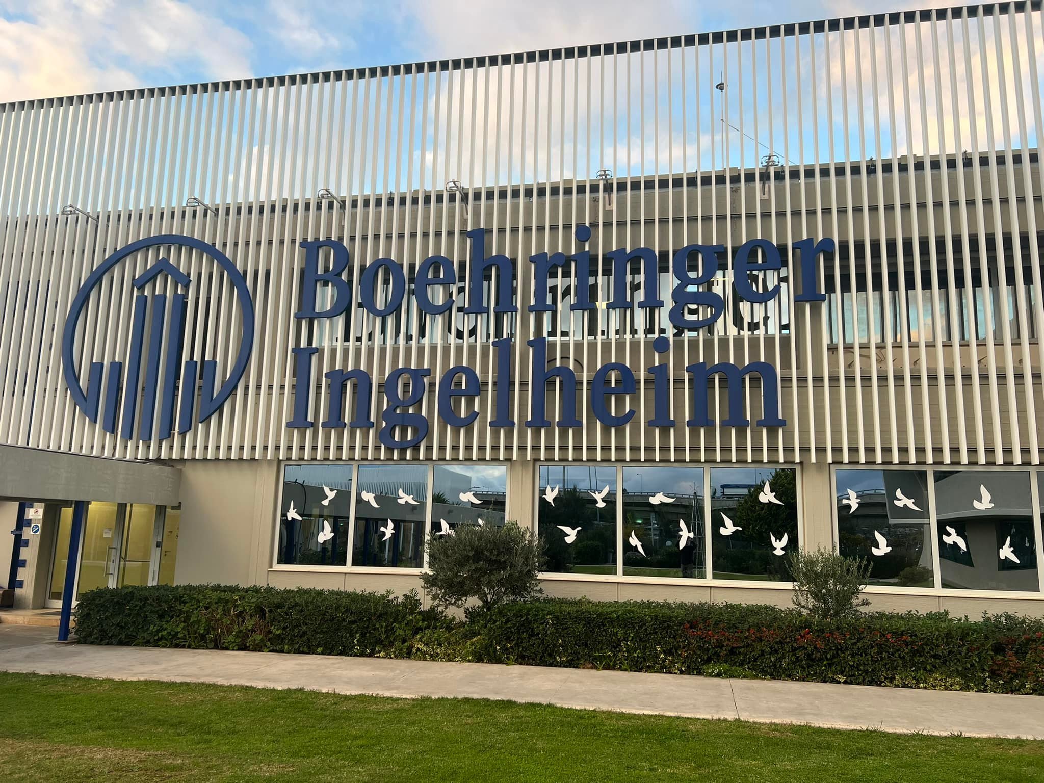 Exterior of Boehringer Ingelheim building with large blue sign and a logo featuring a circle with a stylized upward arrow inside. White bird silhouettes are on the windows below the sign, and there are bushes and a sidewalk in front.