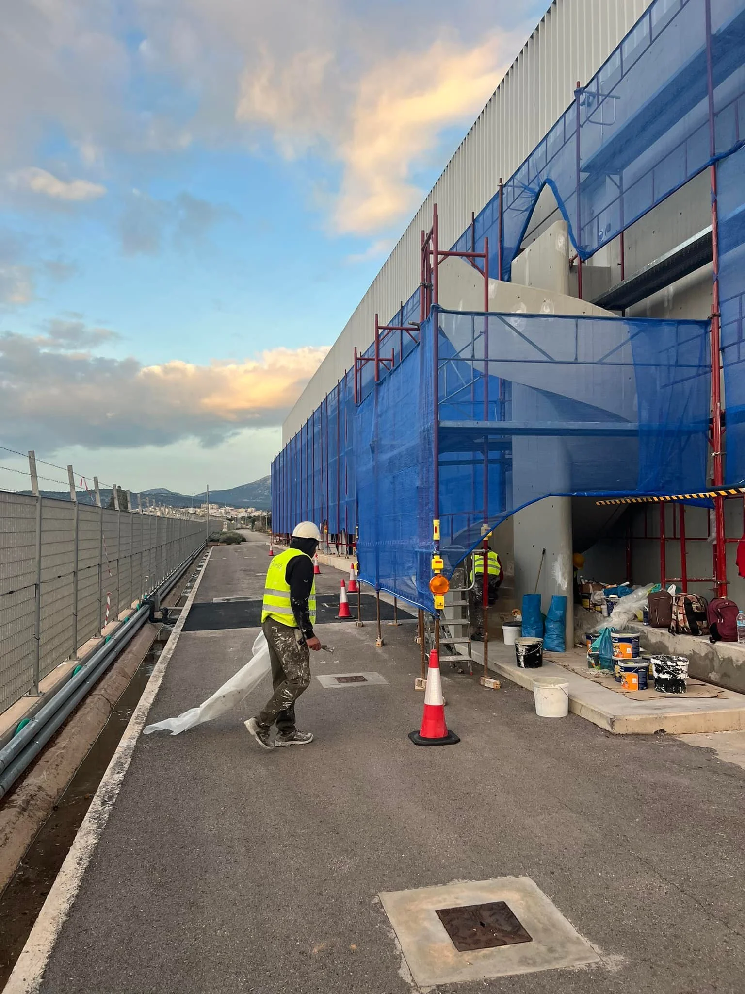 Construction workers in safety vests and helmets working on a building with blue safety netting, orange traffic cones, and construction supplies on the ground, under a partly cloudy sky.