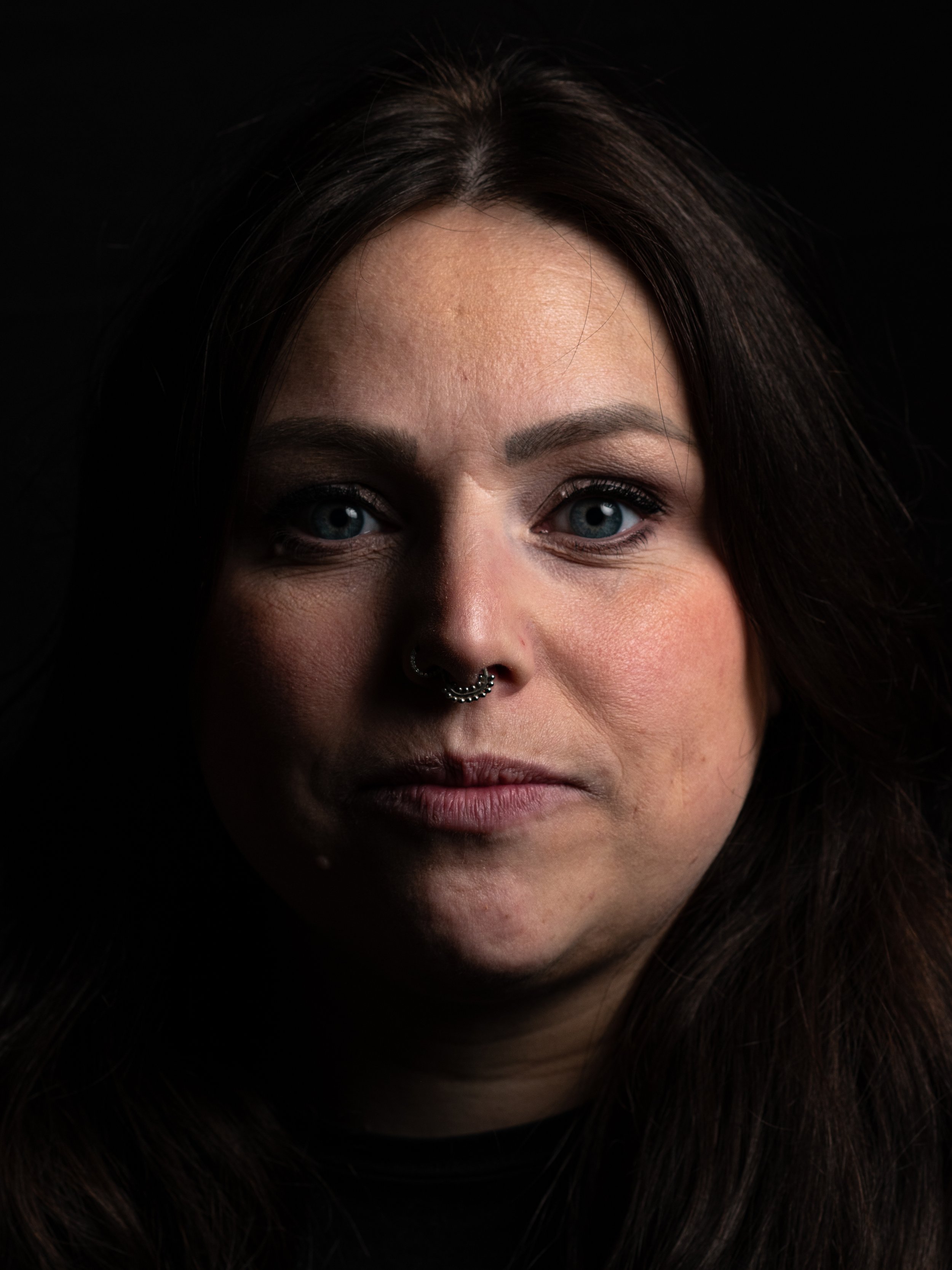 Close-up van een vrouw met blauwe ogen, donkere haar en een neuspiercing, gezicht gedeeltelijk zichtbaar tegen een zwarte achtergrond.
