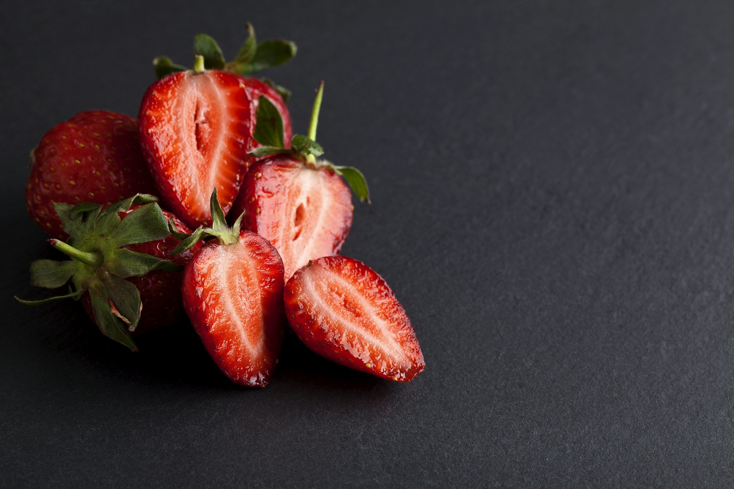 Fresh strawberries, some whole and some sliced, with green leaves, against a dark background.