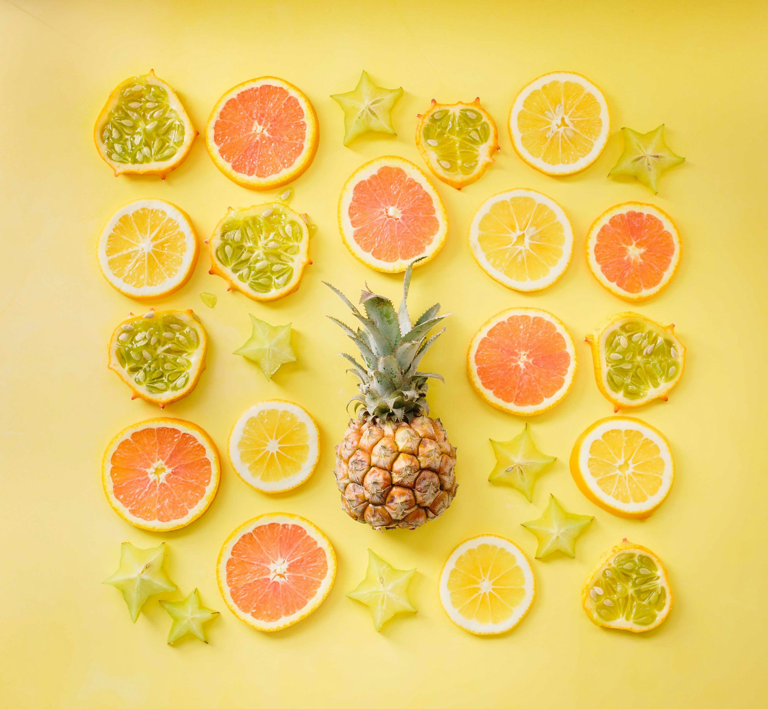 Assorted fruits on a yellow background, including a pineapple, sliced grapefruit, lemon, starfruit, and kiwano melon.