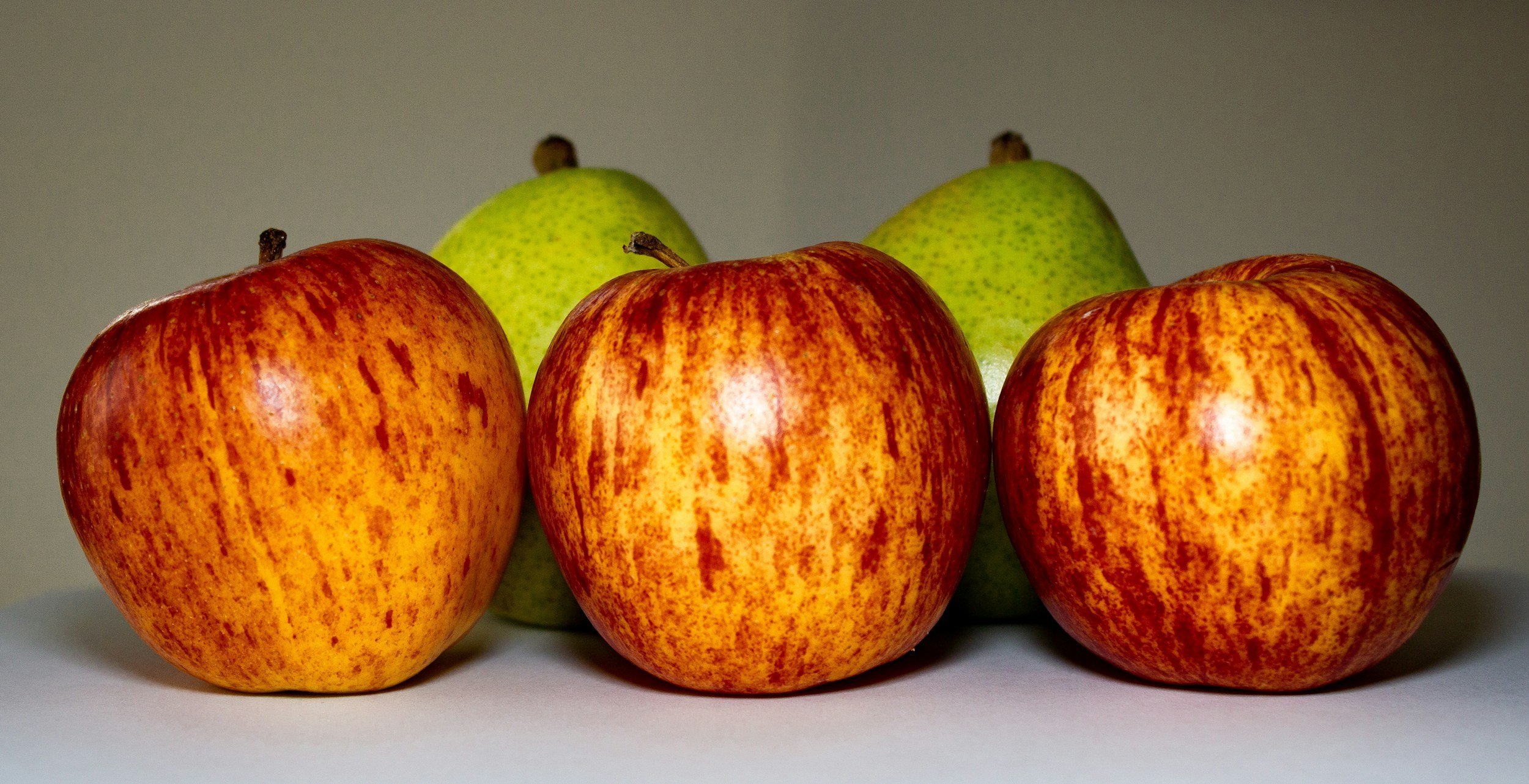 Three red and yellow apples with two green pears in the background on a white surface.