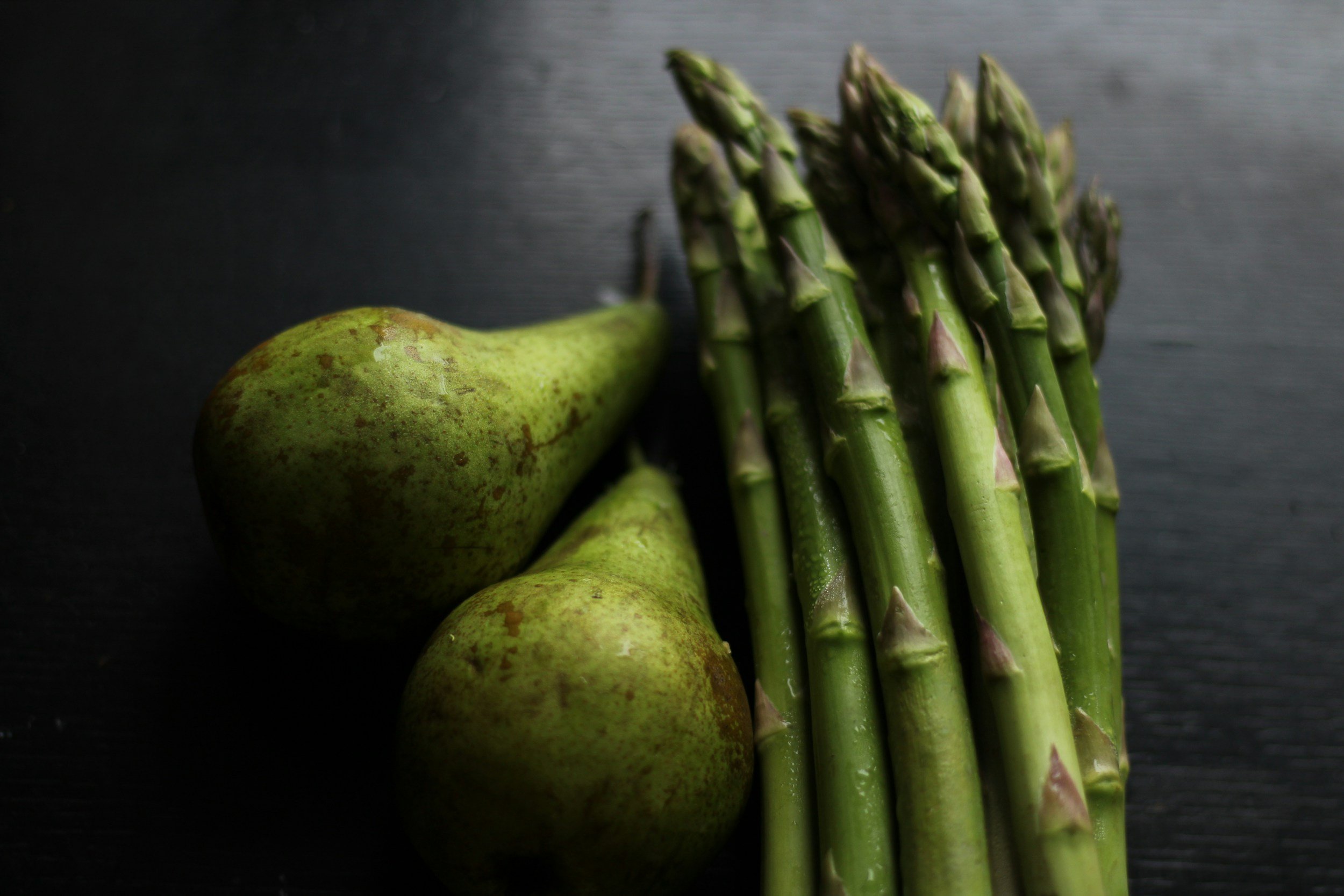 Two green pears next to a bunch of asparagus on a dark surface.