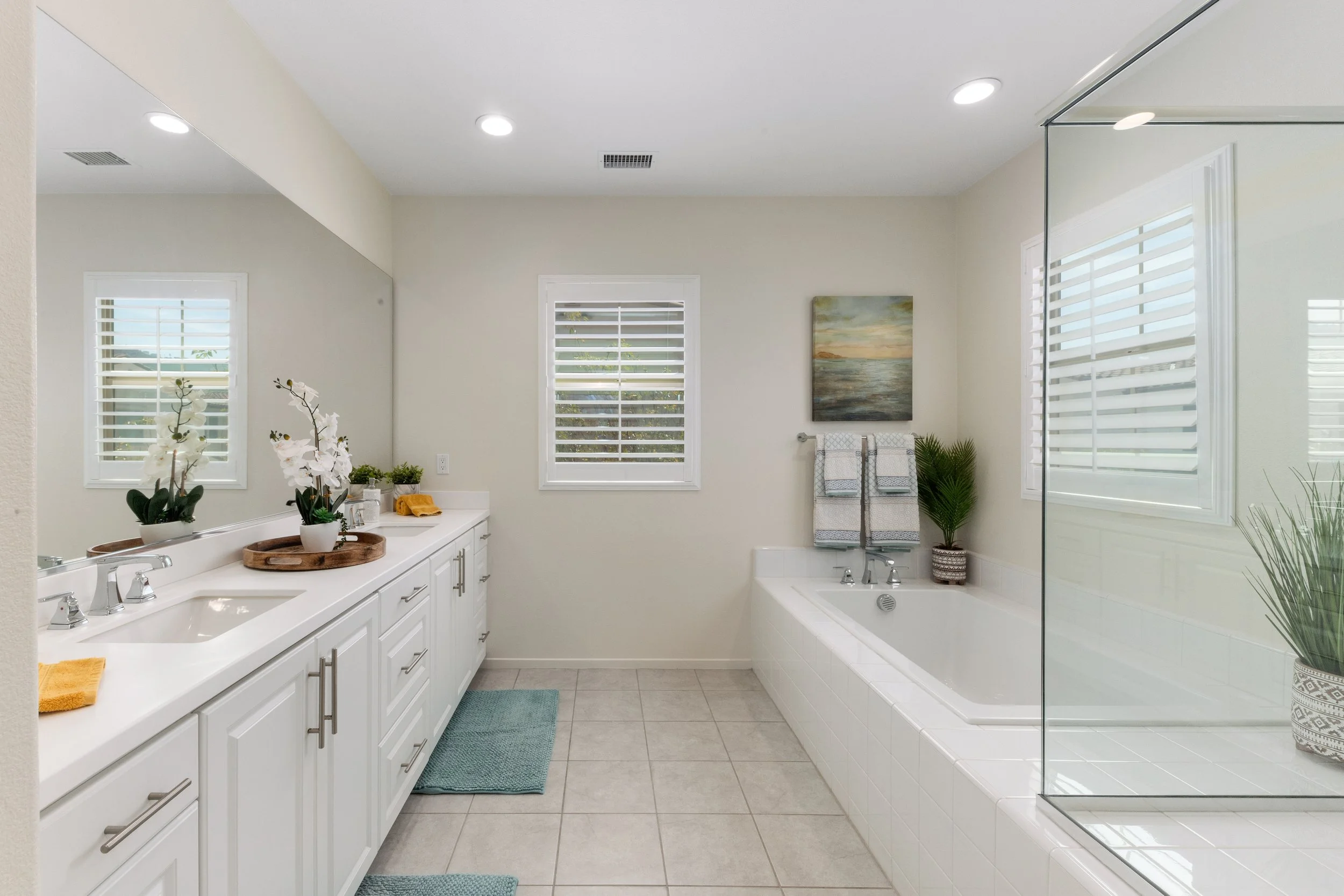 A modern bathroom with white cabinets, a double sink, potted orchids, a large mirror, a bathtub with a painting above, and a glass shower enclosure with a potted plant nearby.