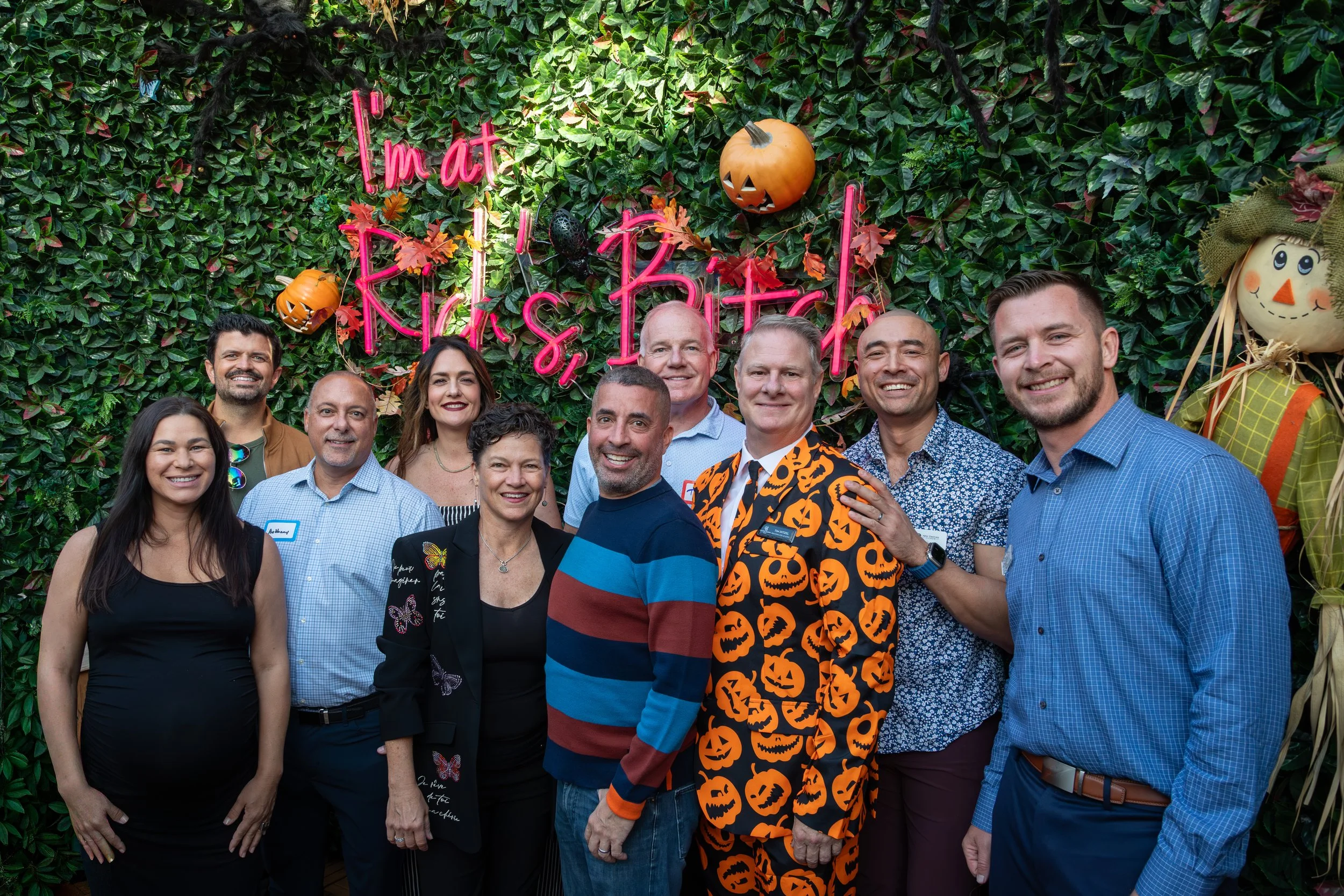 A group of eleven people posing in front of a green leafy wall with Halloween decorations, including carved pumpkins and scarecrows. They are smiling and dressed casually, with one person wearing a Halloween-themed jacket and another wearing a stripe