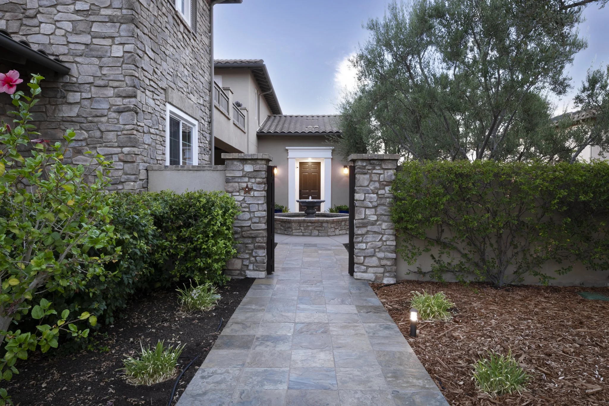 View of a luxury modern house entrance with stone and stucco exterior walls, a tiled roof, a small pathway with stone tiles, surrounded by greenery, with lighting fixtures on the wall and small landscape lights on the ground.