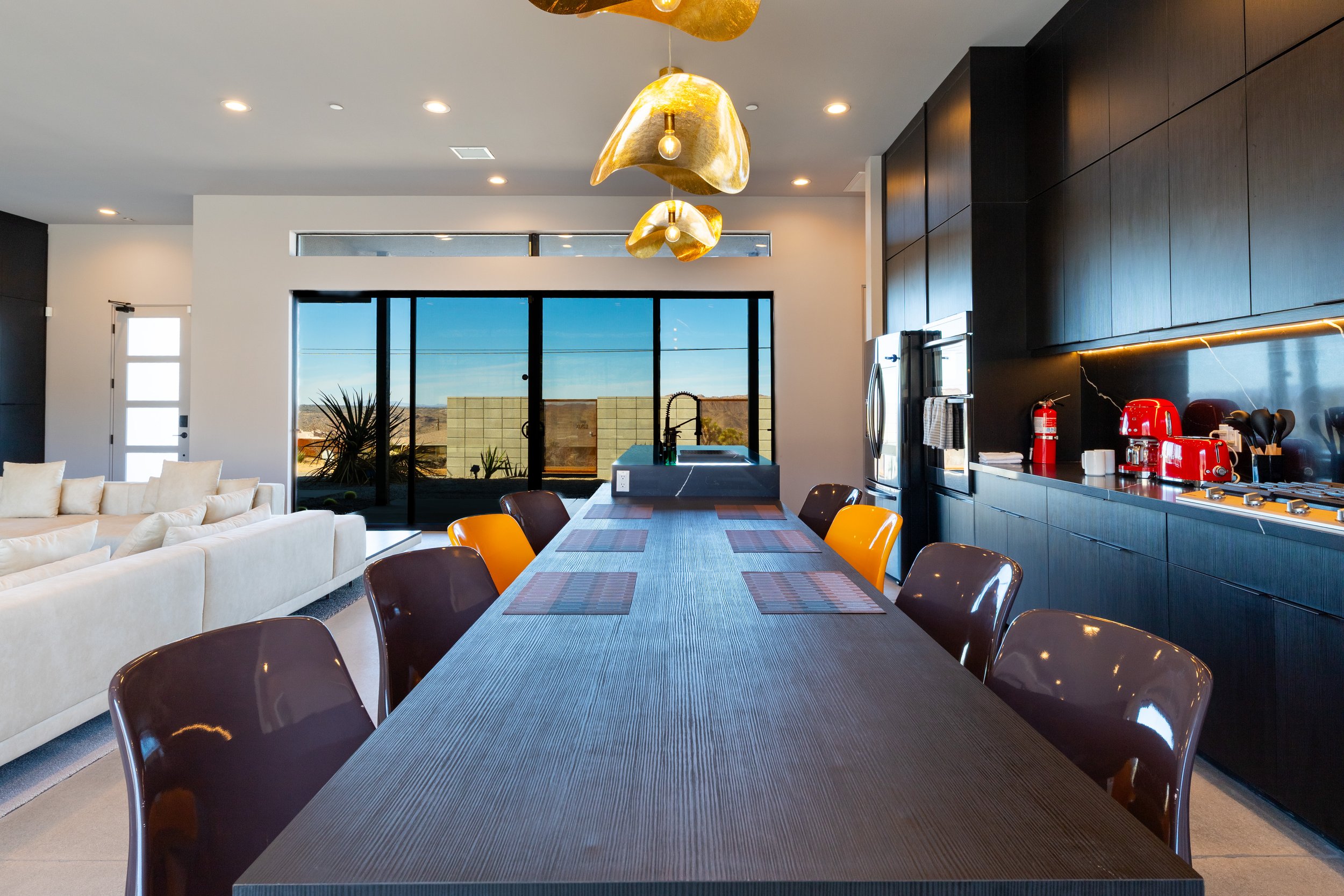Modern open-plan kitchen and dining area featuring a long dining table with placemats, black and orange chairs, a kitchen with dark cabinetry, a stainless steel refrigerator, bright hanging pendant lights, large windows showcasing a scenic landscape 