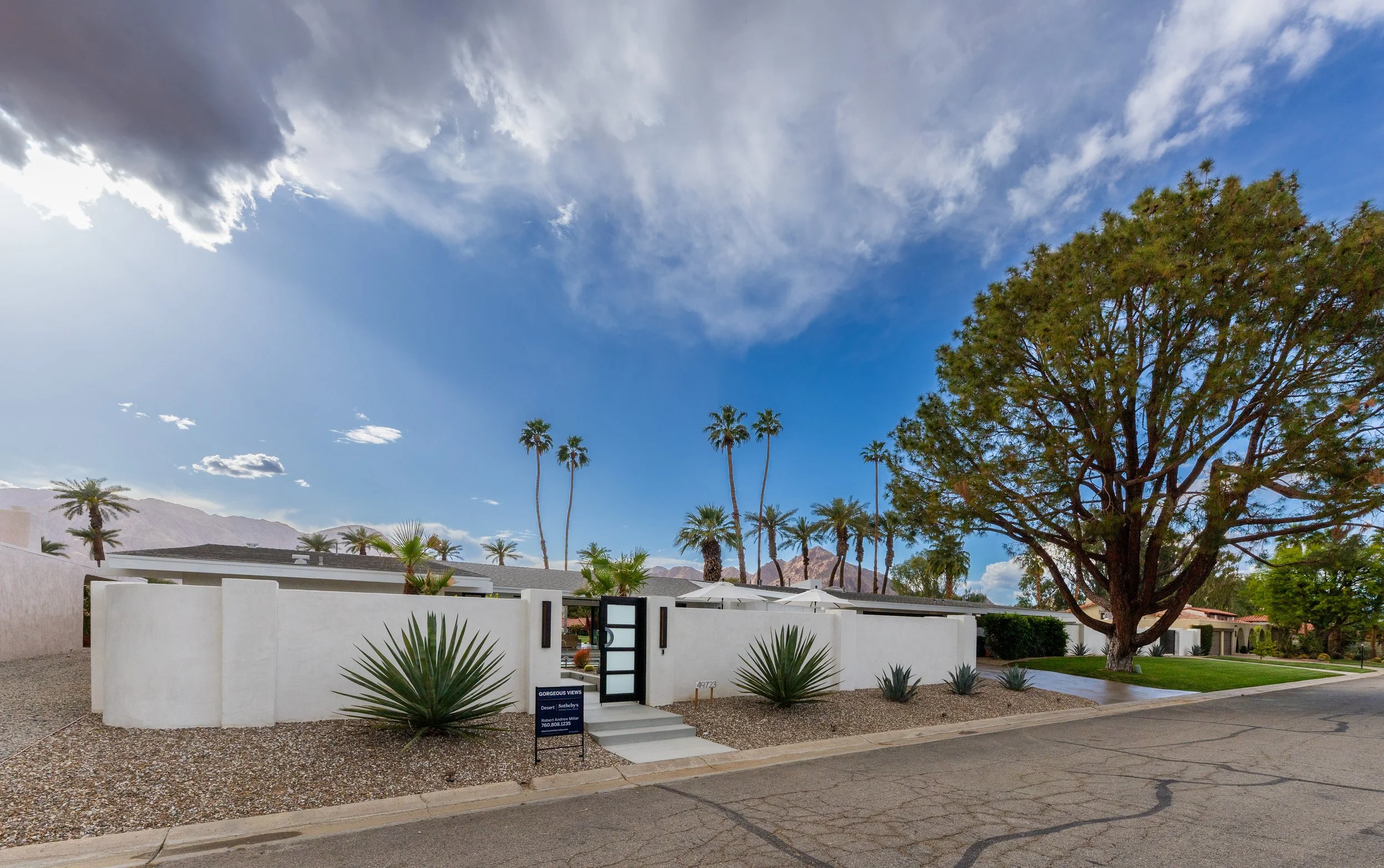 A modern house with white exterior walls and a flat roof, surrounded by desert landscaping, with tall palm trees and a large tree in the front yard. The sky is partly cloudy with sun rays peeking through.