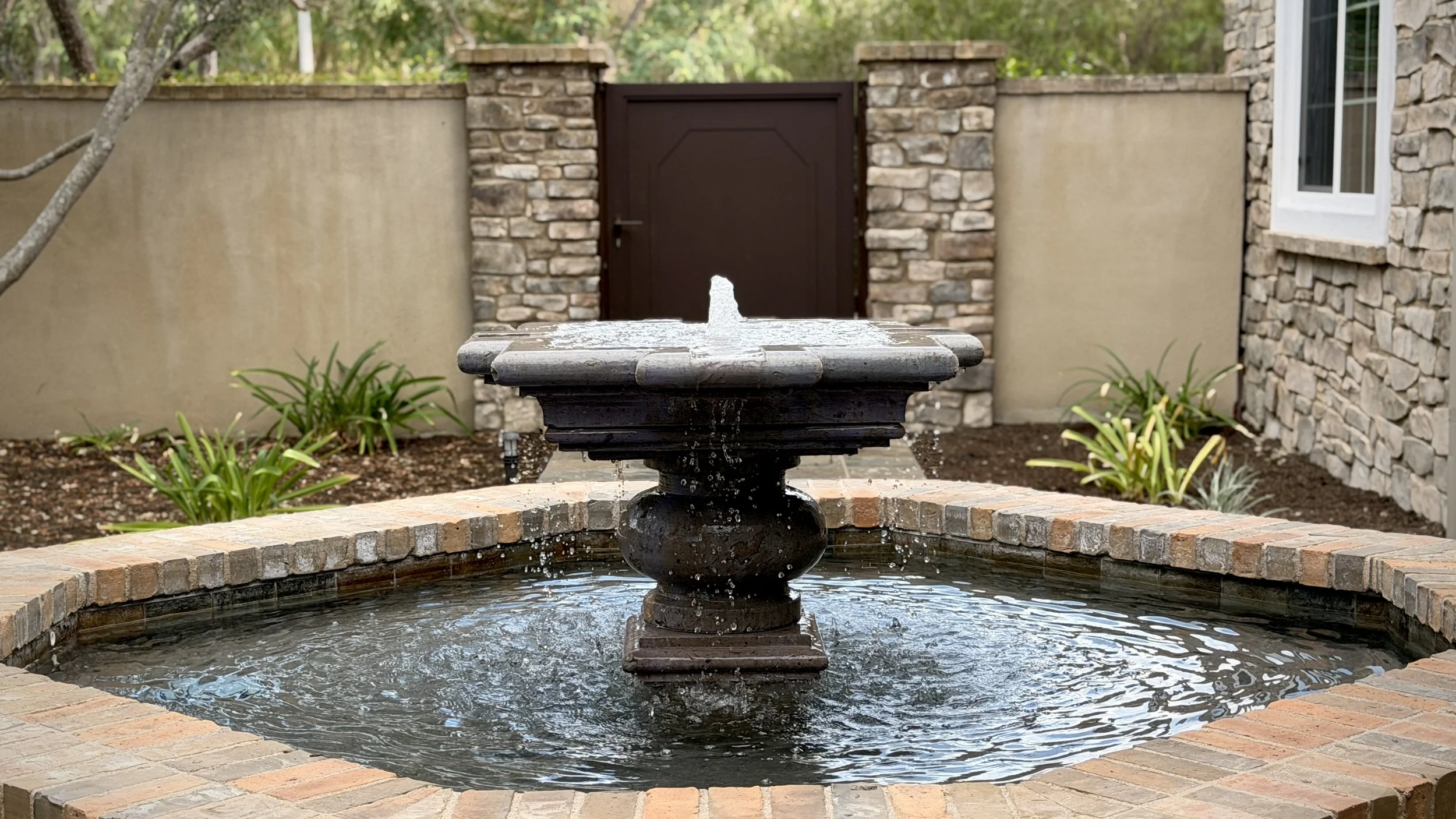 A decorative fountain in a courtyard surrounded by brick, with plants and a stone wall in the background.