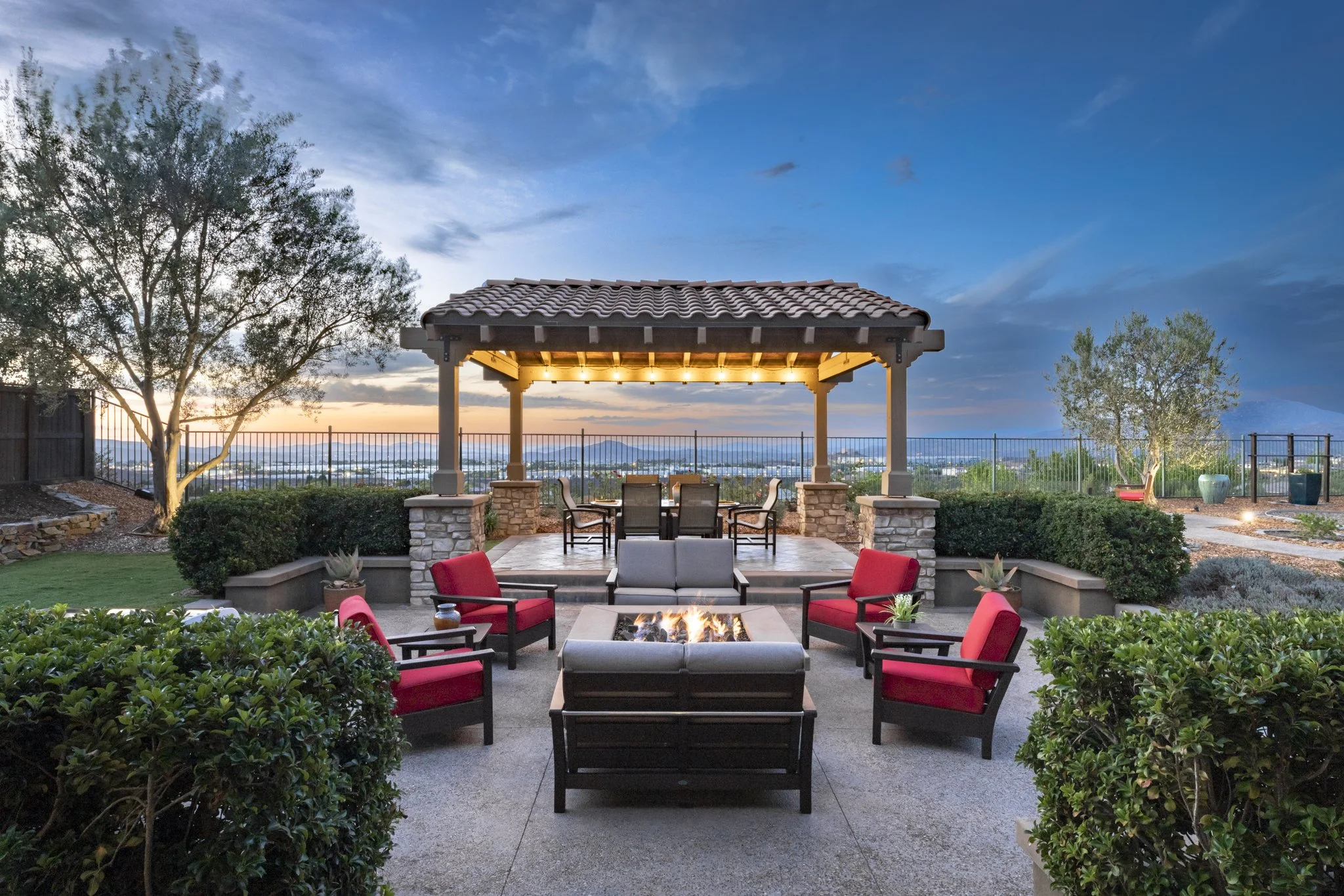A multi-million dollar property outdoor patio area at dusk with a fire pit, comfortable seating, a gazebo with lights, trees, and a view of the city skyline and mountains.