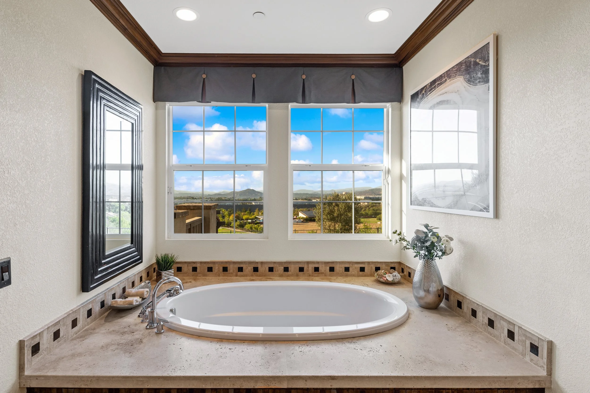 Bathroom with bathtub near two large windows showing a view of blue sky and mountains, decorated with a large mirror, potted plant, and decorative stones.
