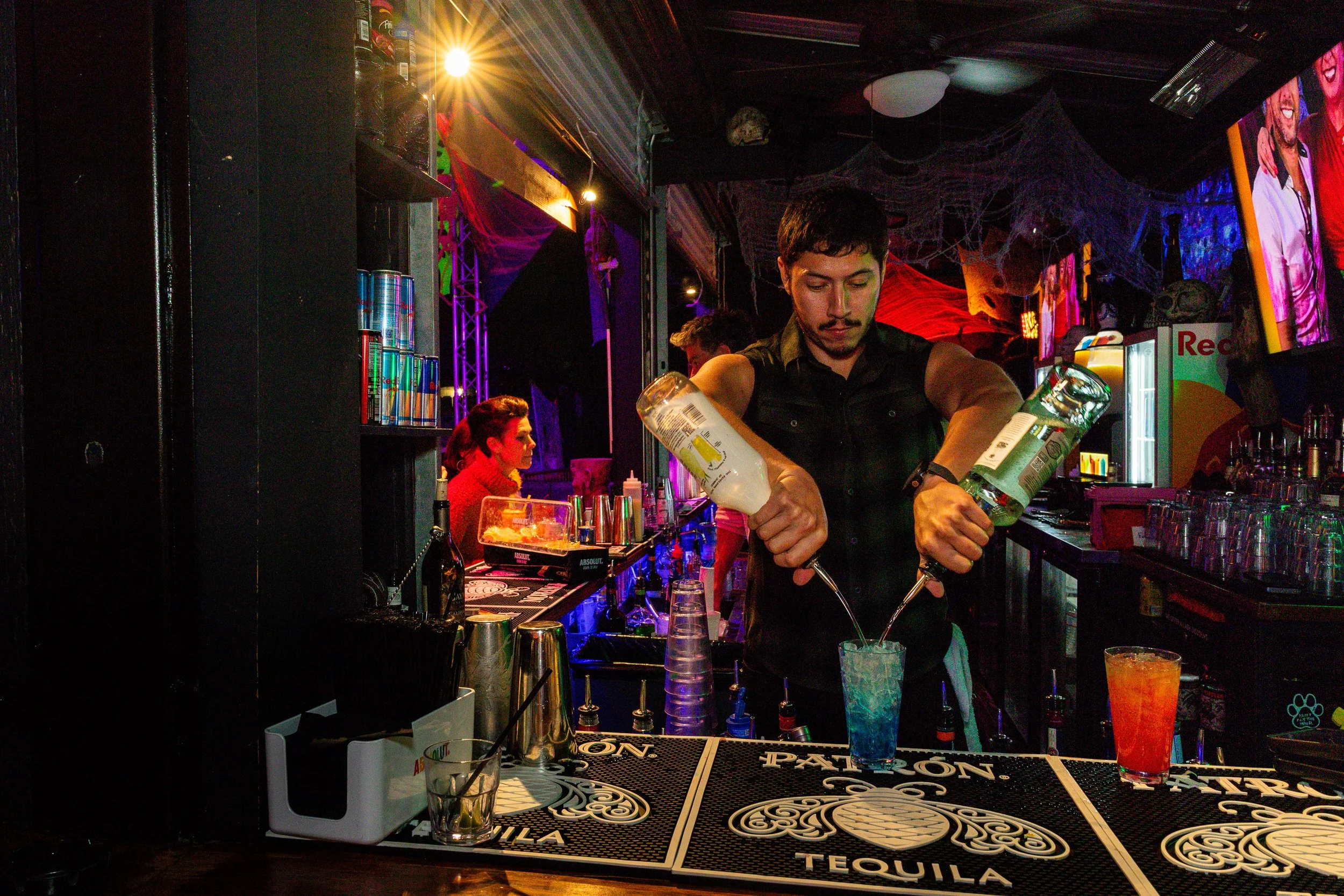 A bartender is pouring drinks in a nightclub with colorful lighting, neon signs, and a decorated bar counter with various bottles, mixers, and glasses. A woman with red hair is seated at the bar.