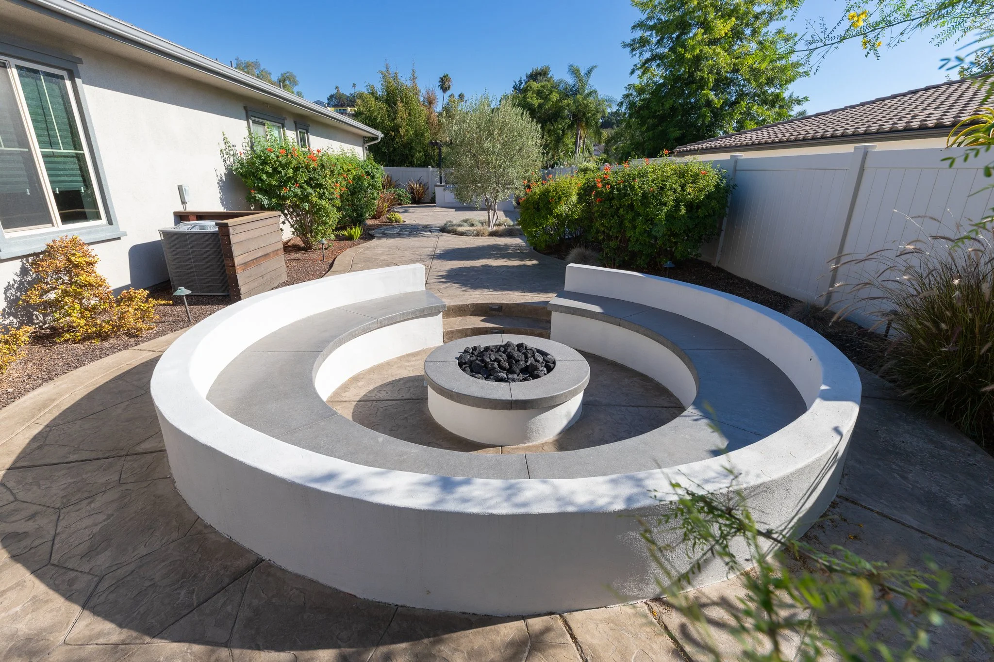 A luxurious backyard patio with a circular built-in fire pit in the center, surrounded by curved seating, with various green shrubs, trees, and a white fence under a blue sky.