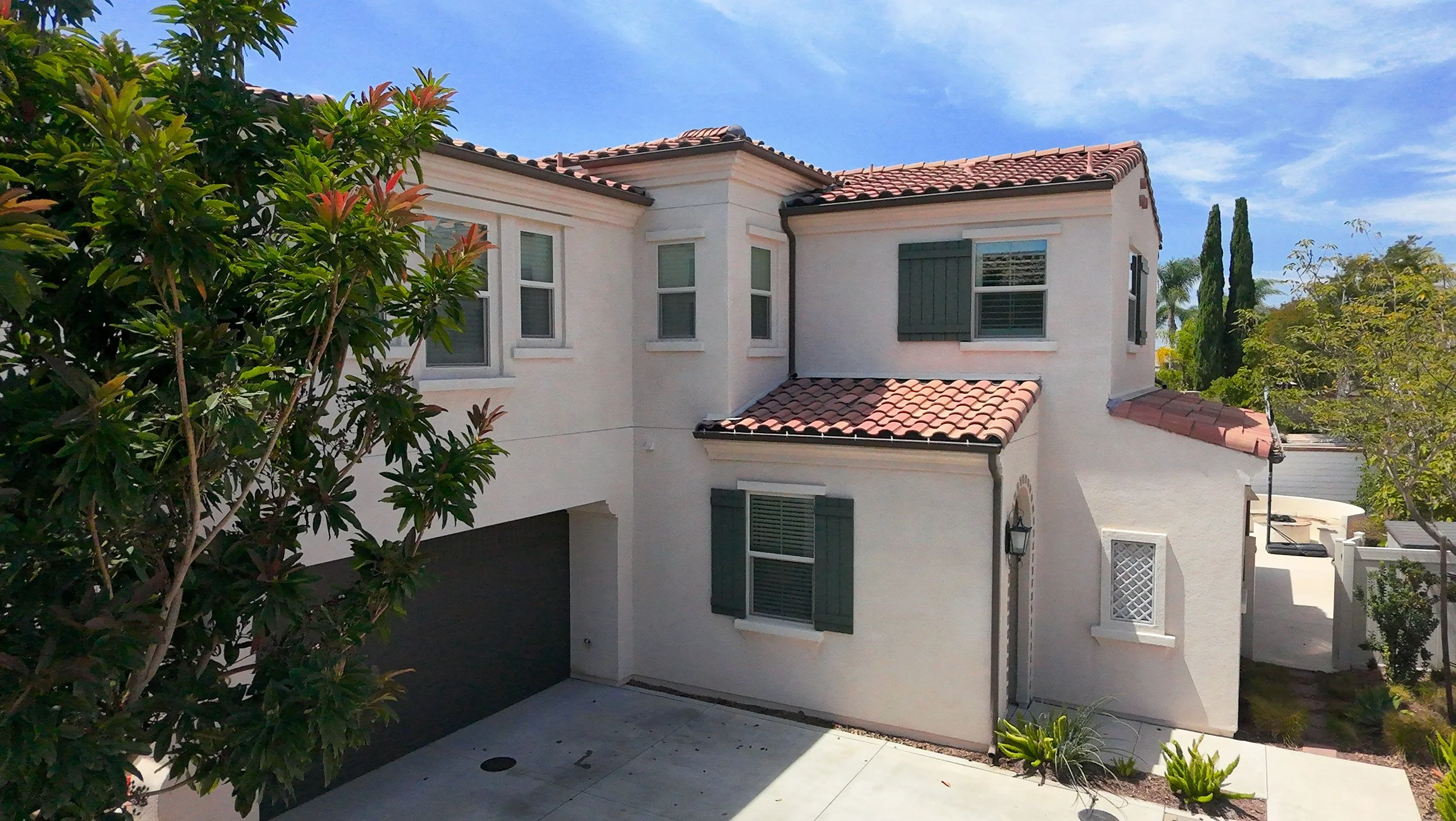 A two-story house with white stucco walls, dark green window shutters, and a red tile roof is visible. The house is surrounded by trees and bushes, with a concrete patio in the foreground and a clear blue sky overhead.