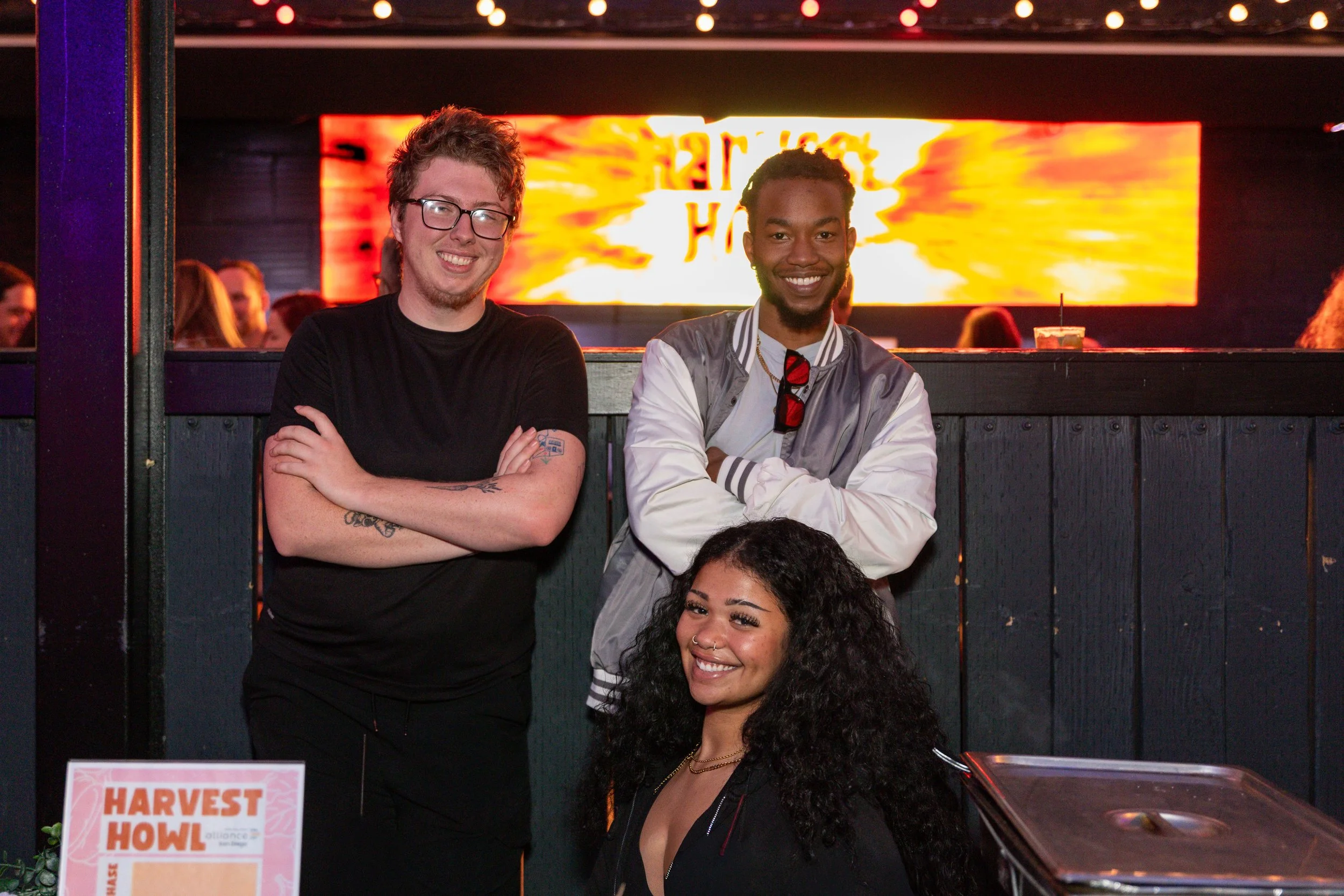 Two young men and a young woman smiling at the camera, standing together at a social event. The background is illuminated with a bright, colorful digital display.