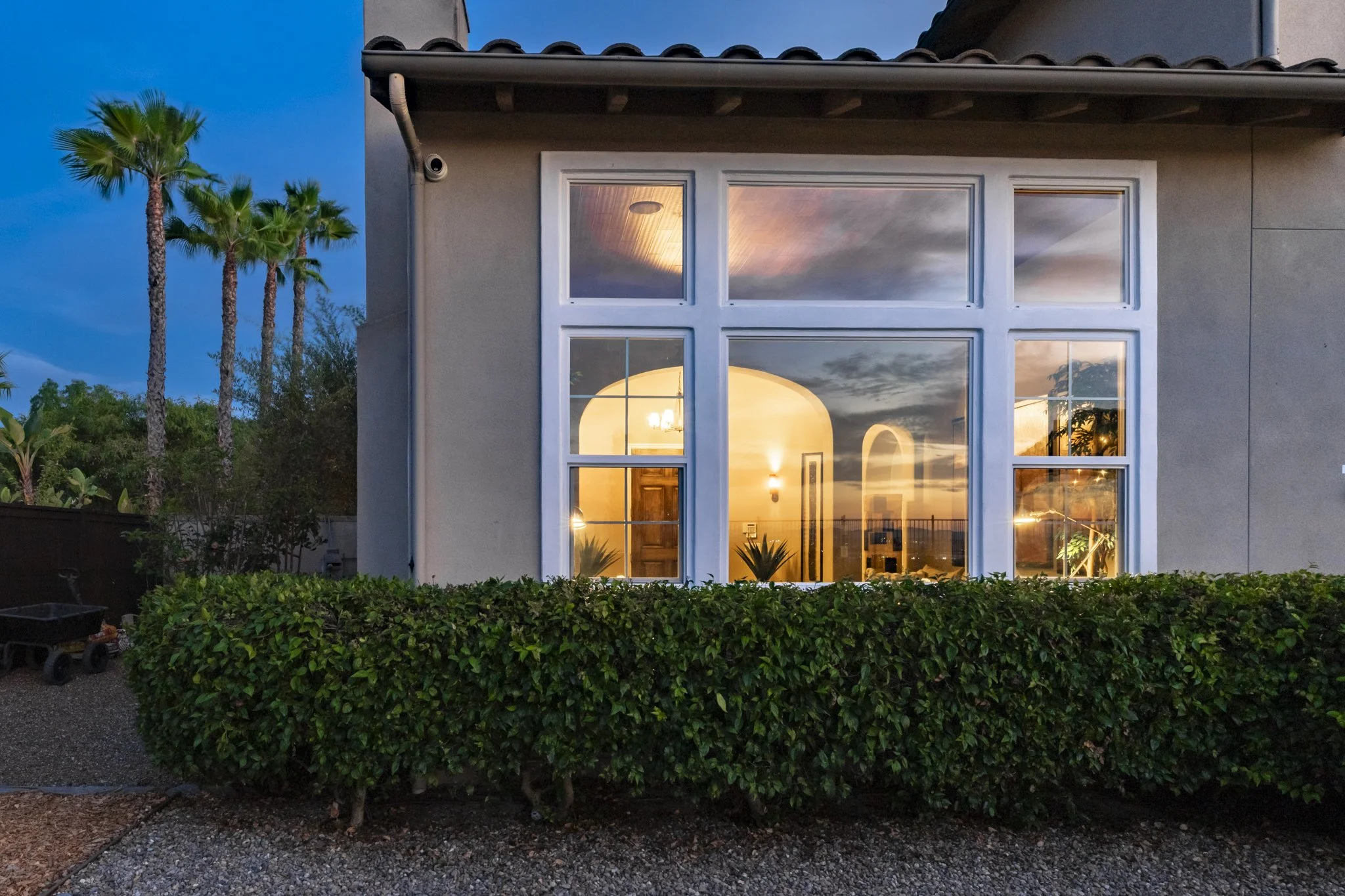 View of a luxury house with large windows illuminating the interior, outdoor garden bushes in the foreground, and palm trees against a blue twilight evening sky.