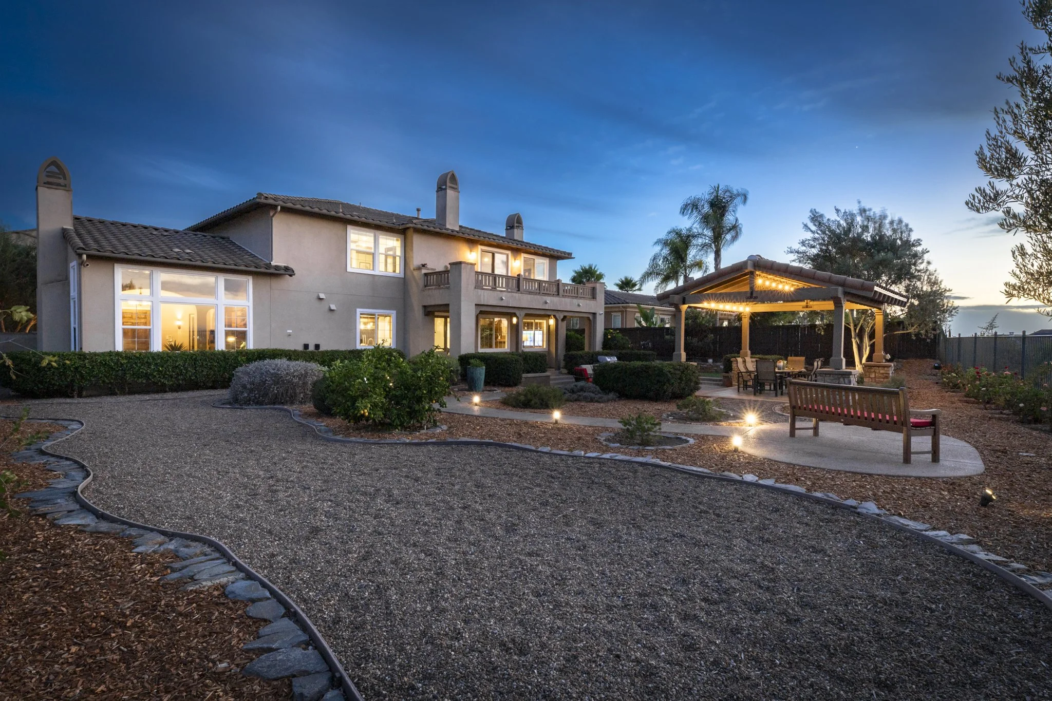 A large luxury San Diego two-story house with large windows and a balcony in a landscaped backyard at dusk, illuminated by outdoor lighting, with a patio area covered by a pergola, surrounded by trees and shrubbery.