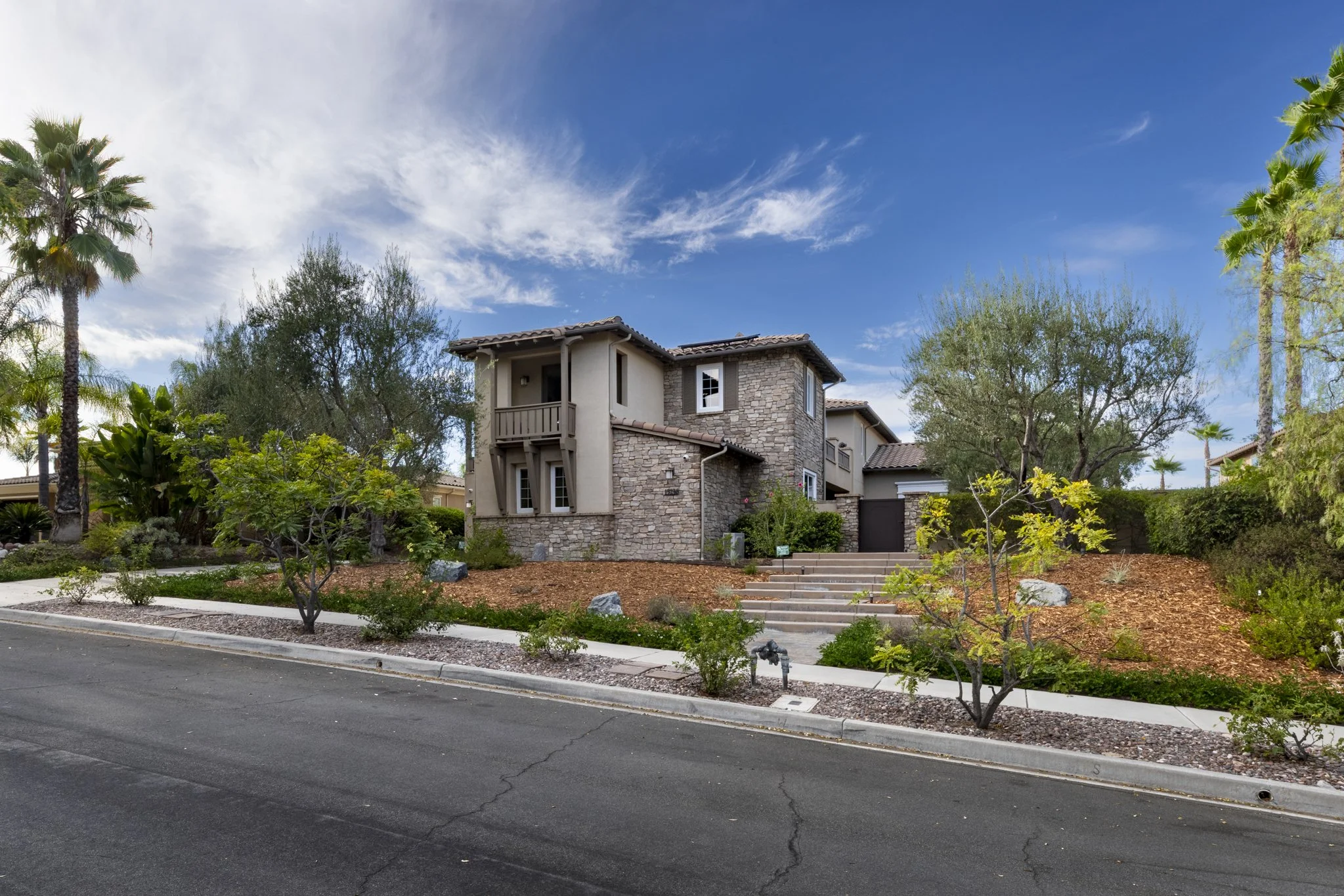 A multi-million dollar two-story house with a stone and stucco exterior, surrounded by trees and shrubs, with a clear blue sky above.