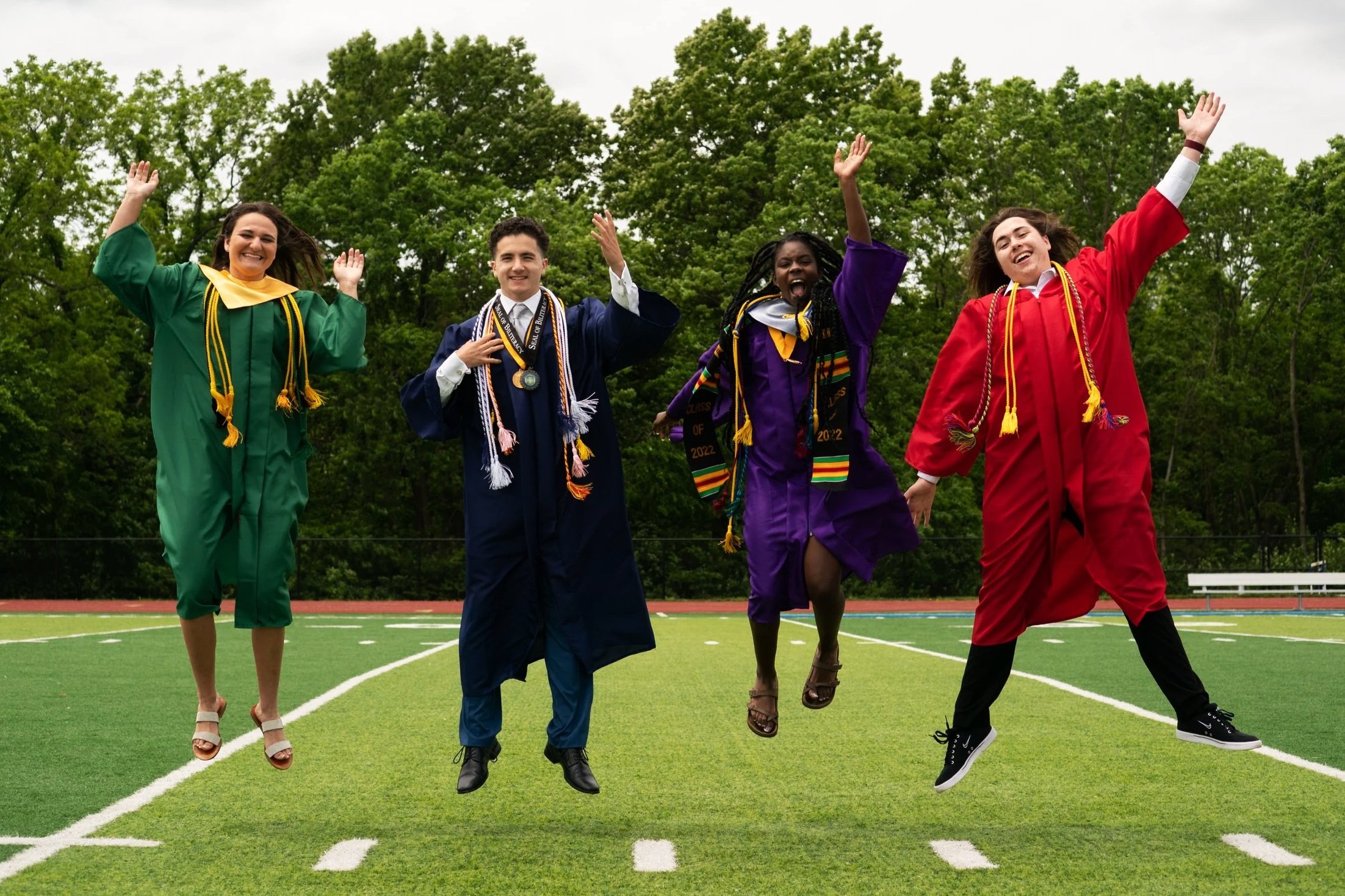 Four NKC Schools graduates in caps and gowns jump on a football field.