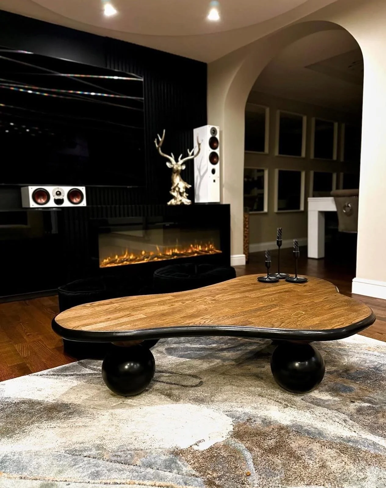 Modern living room with a wooden coffee table, featuring 3 black ball legs, a fireplace, decorative items on the mantle, and a TV mounted on the wall.