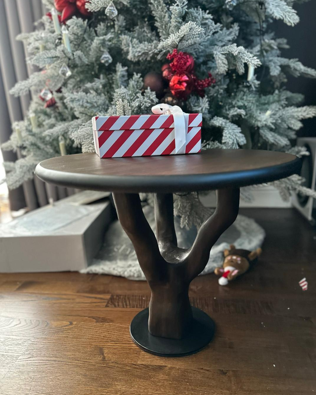 A Christmas scene with a decorated Christmas tree in the background and a small gift box wrapped with red and white striped paper and a white ribbon on a wooden side table in the foreground.