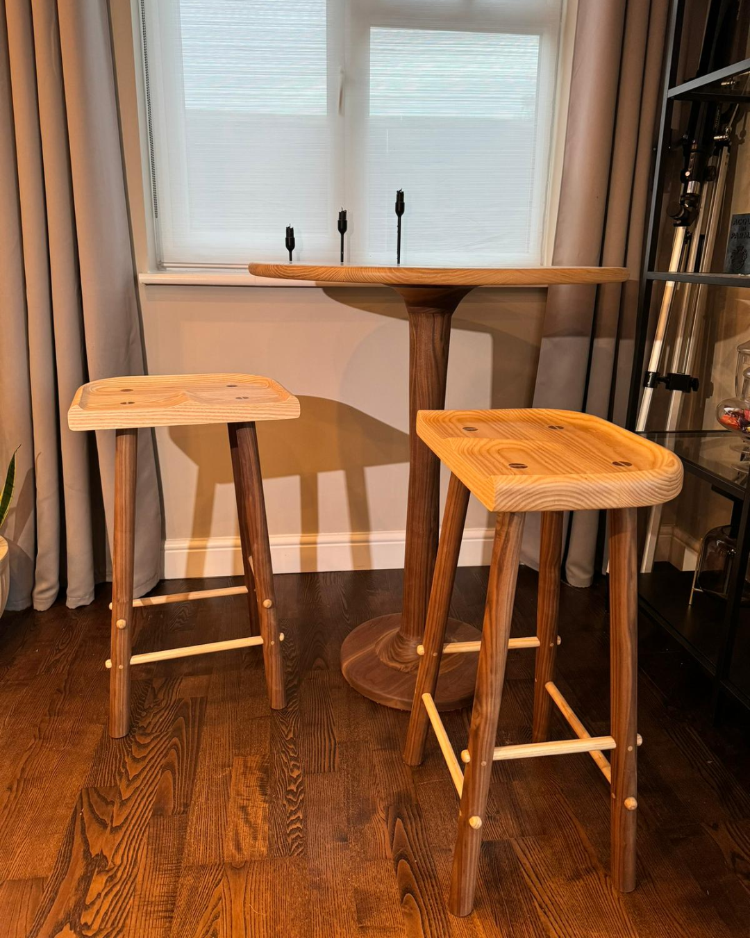 Wooden high bar table with two matching wooden stools, placed on hardwood floor near window with blinds and curtains.