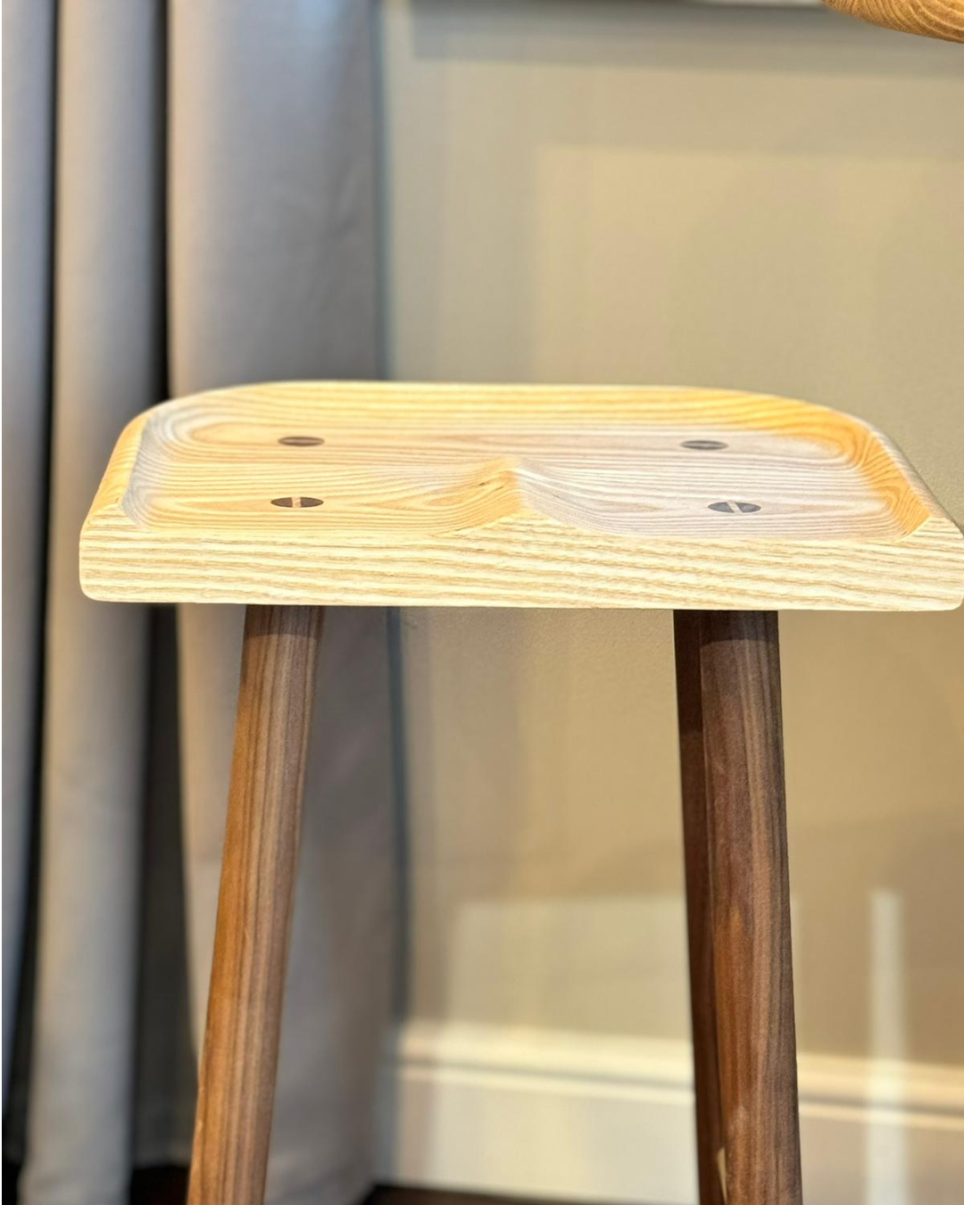 A wooden stool with a square, slightly recessed seat featuring screw holes, standing on three angled legs near a wall.