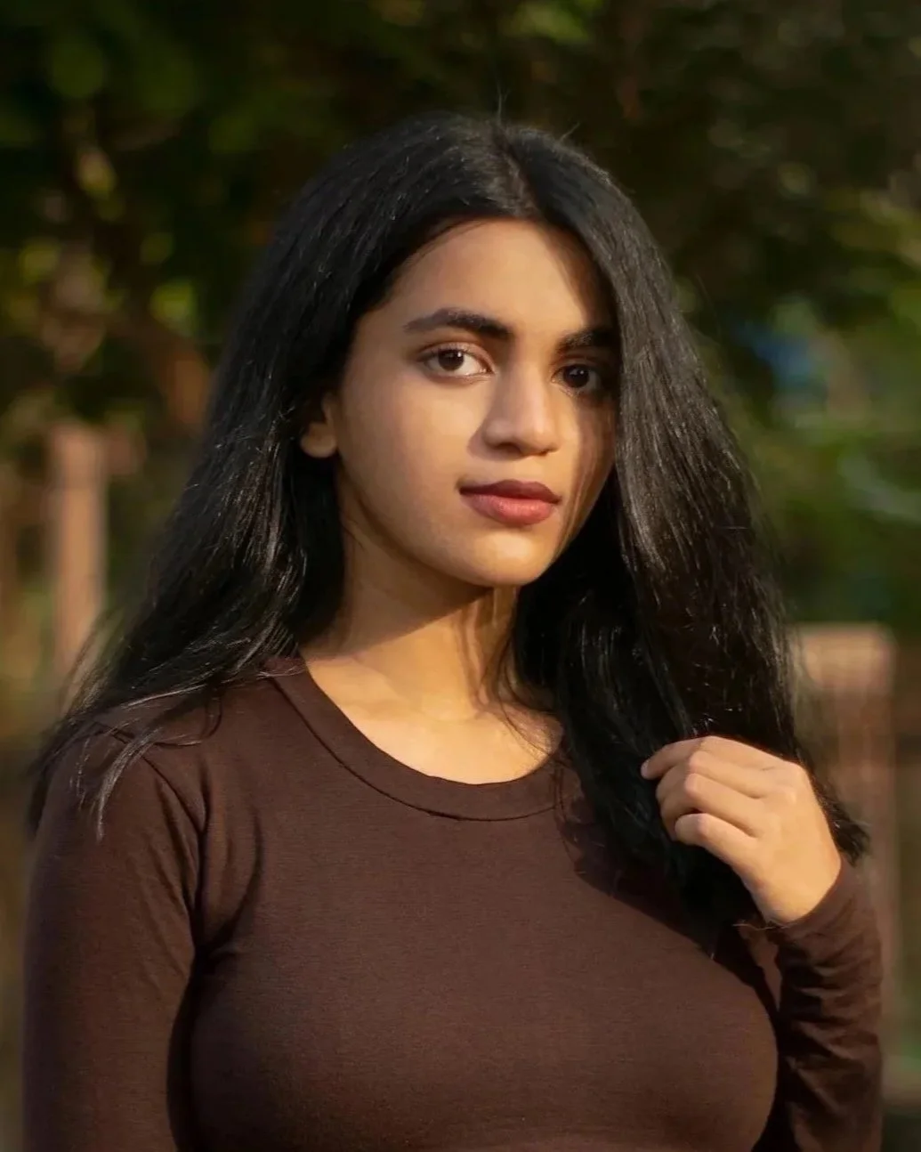 A young woman with long dark hair wearing a white shirt and black necklace stands on a balcony with metal railings overlooking a cityscape with a tall building in the distance. She has her eyes closed, appears relaxed on an overcast day.