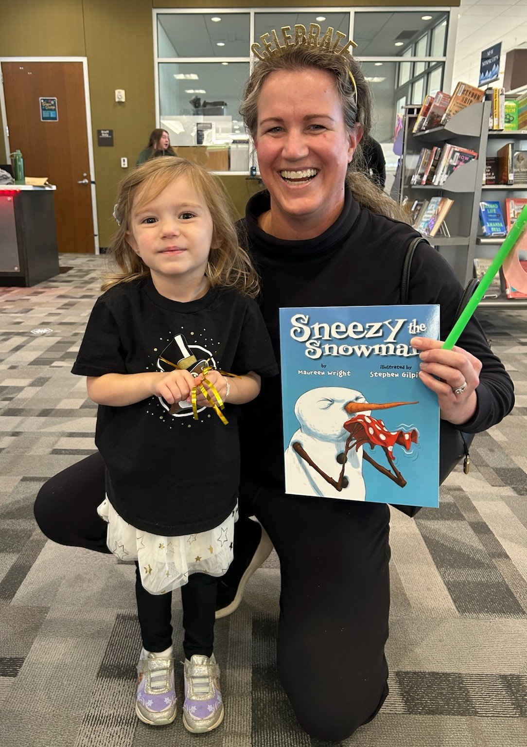 A woman and a young girl are smiling for a photo in a library. The woman is holding a children's book titled 'Sneezy the Snowman' and a green straw, while the girl is holding a yellow ribbon. The woman has a 'Celebrate' headband, and the girl is wearing a black shirt with a space-themed design and a white skirt with stars.