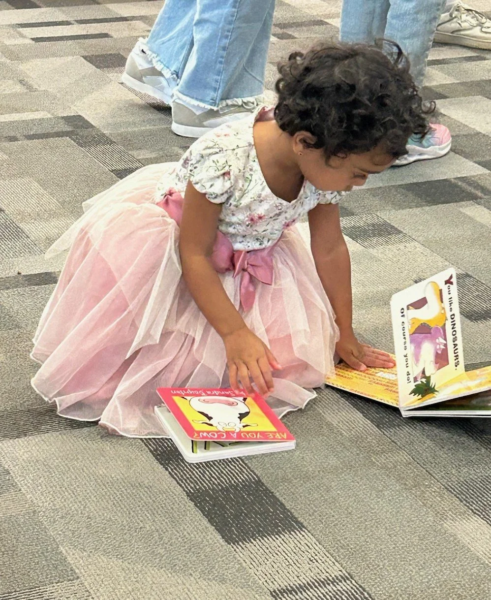 A young girl with curly hair wearing a floral shirt and pink tutu skirt is sitting on a carpeted floor, reading colorful children's books about dinosaurs.