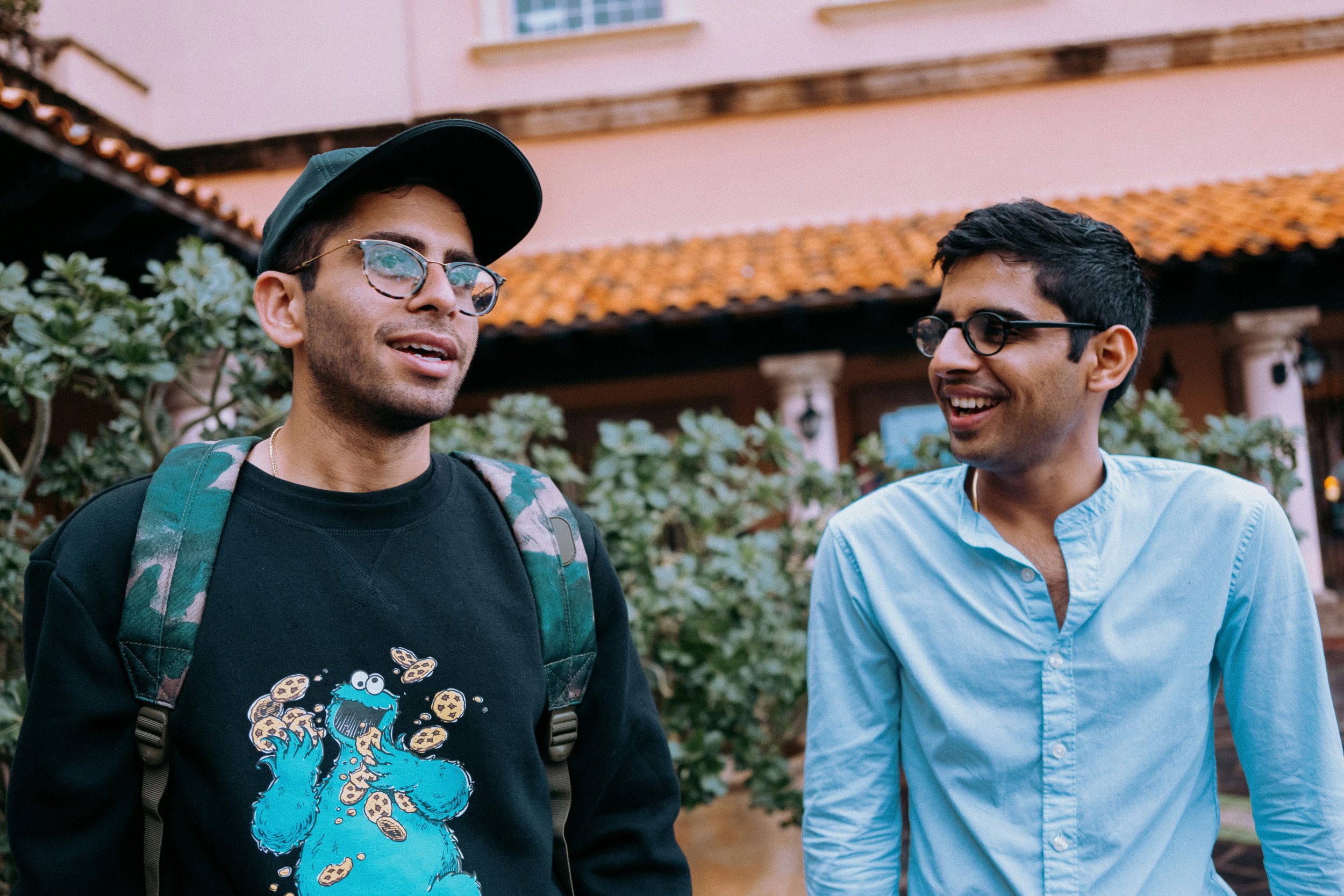 A vibrant image of South Asian male siblings, both older adolescents, enjoying a moment of conversation, smiles, and laughter outdoors. They are standing together, with one sibling wearing a backpack, suggesting they are traveling or on an adventure.