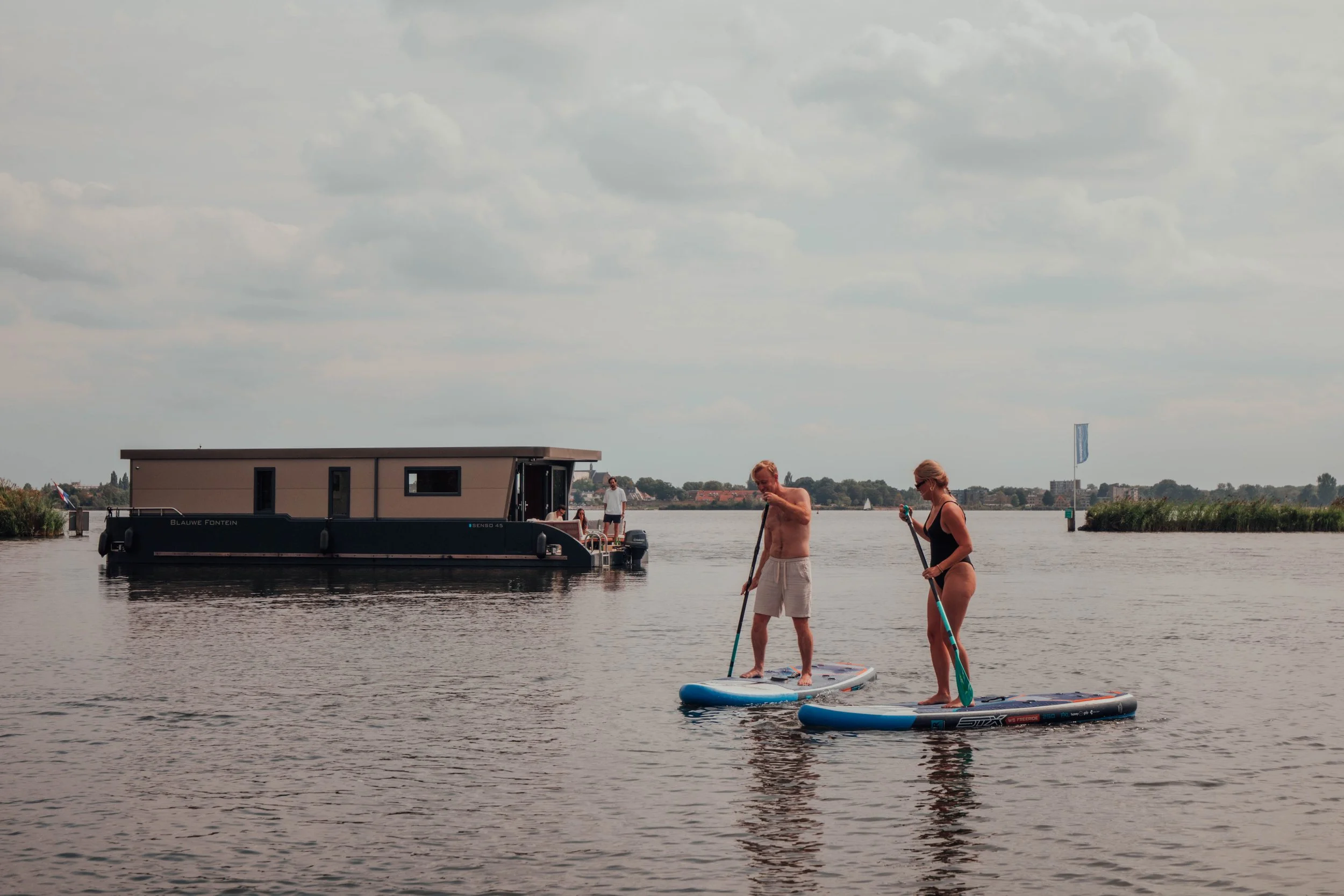 Two people paddleboarding on a lake with a houseboat in the background under a cloudy sky.