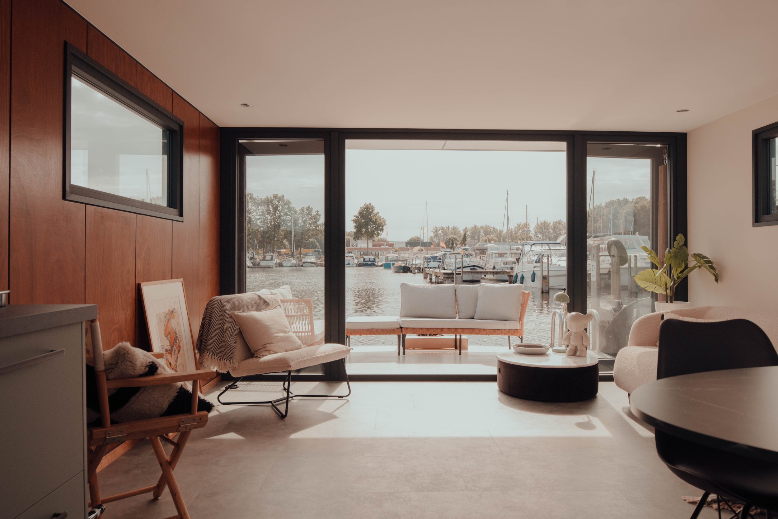 Interior of a modern living room with wooden walls and large windows overlooking a marina. The room features a sofa, chairs, a circular coffee table, and a small potted plant. The outdoor view includes boats and water.