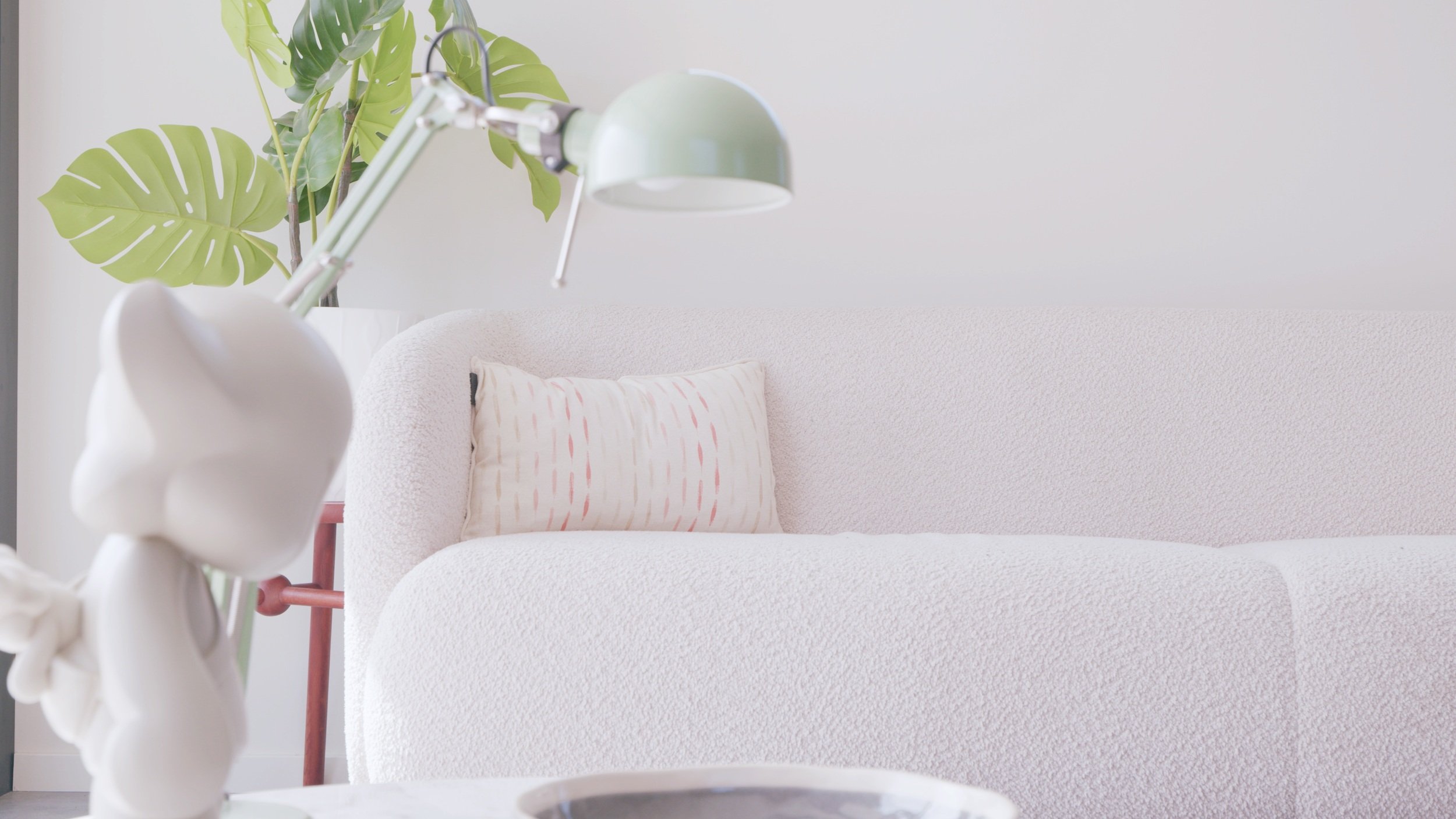 Minimalist living room with a white textured sofa, decorative pillow, green desk lamp, and tropical plant in the background.