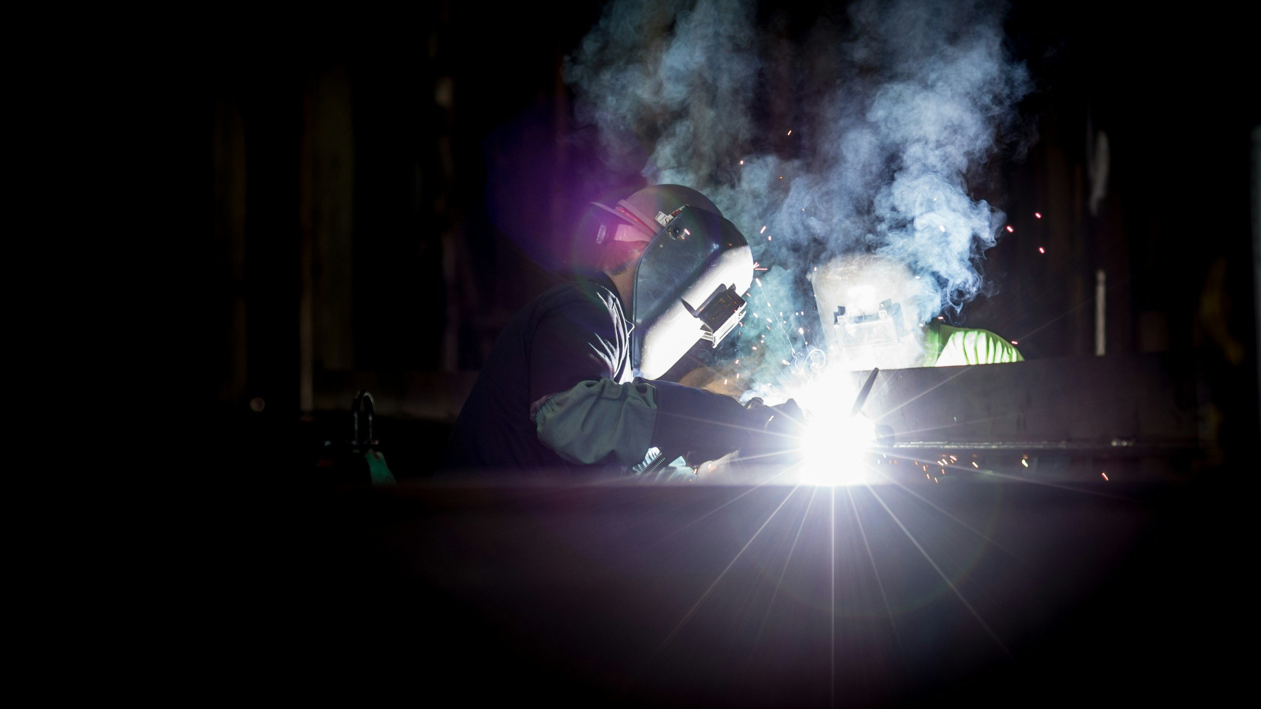 Welder working with bright sparks and smoke in a dark industrial setting.