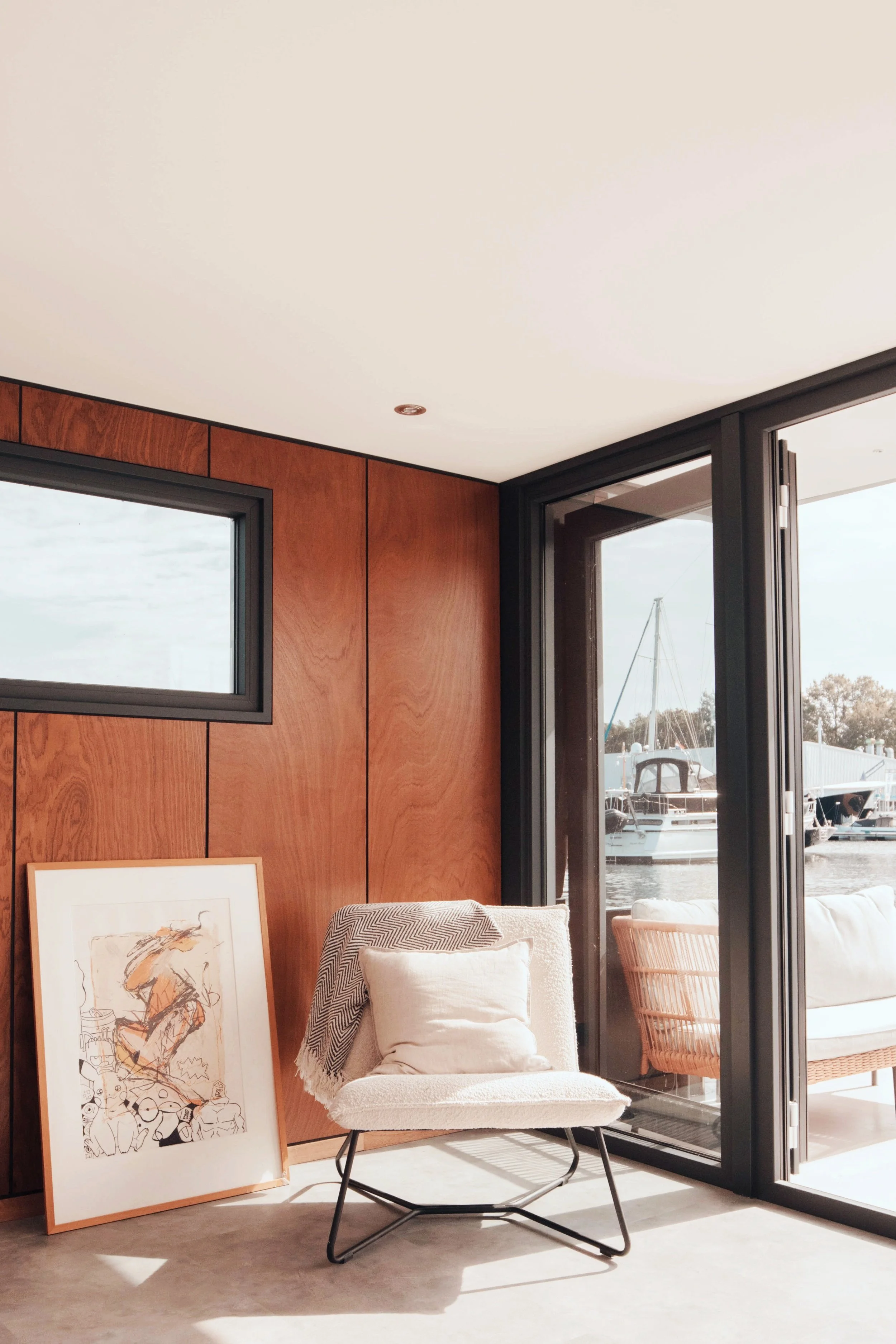 Cozy indoor nook with a chair, decorative pillows, a wooden wall art piece, and large glass doors overlooking a marina.