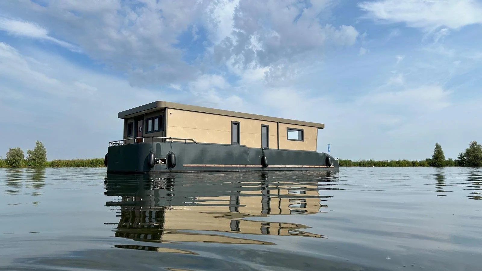 Modern houseboat on a calm lake with a clear blue sky and some trees in the background.