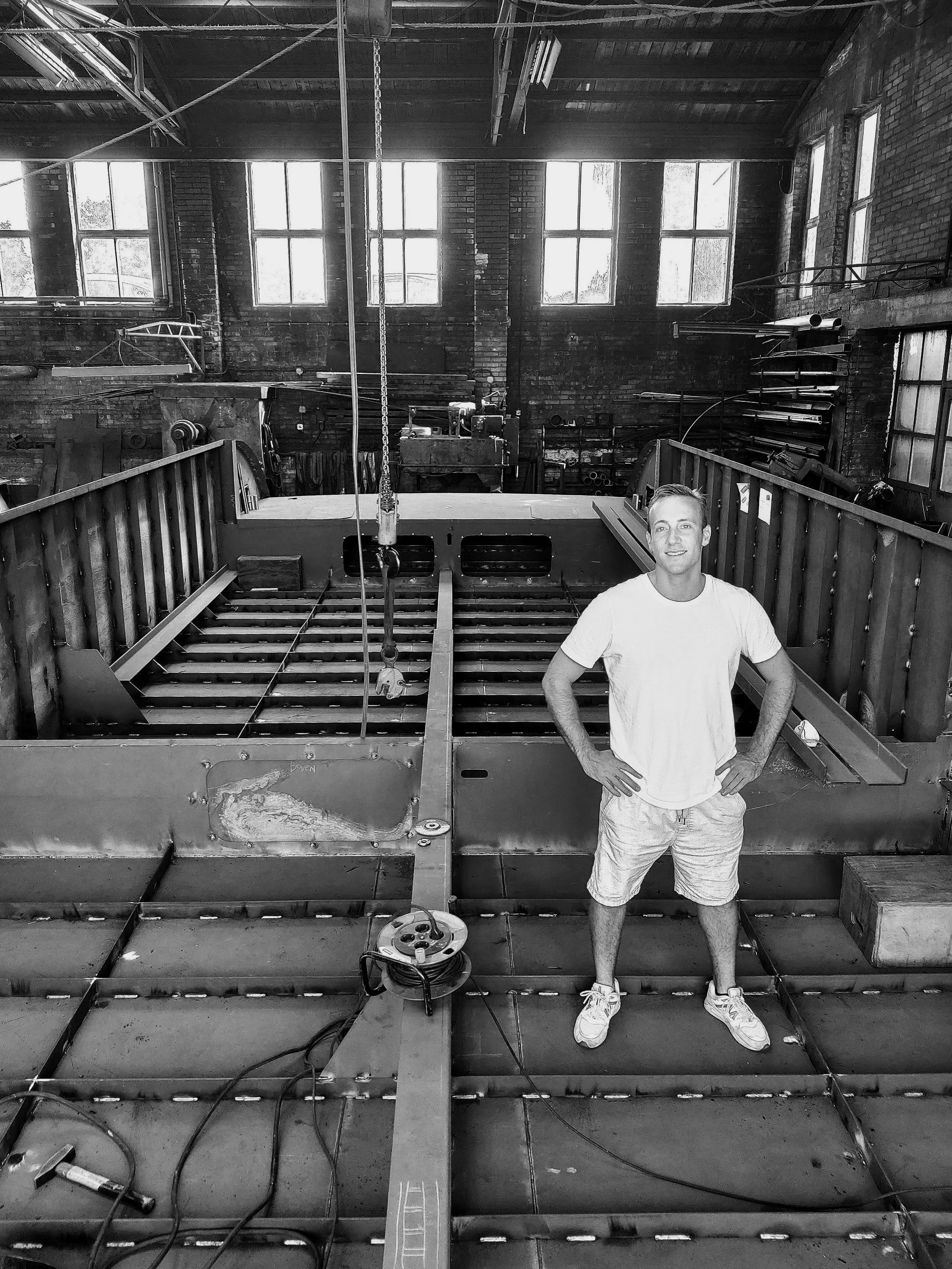 Black and white image of a man standing in a large industrial workshop, with metal beams and construction materials around him.