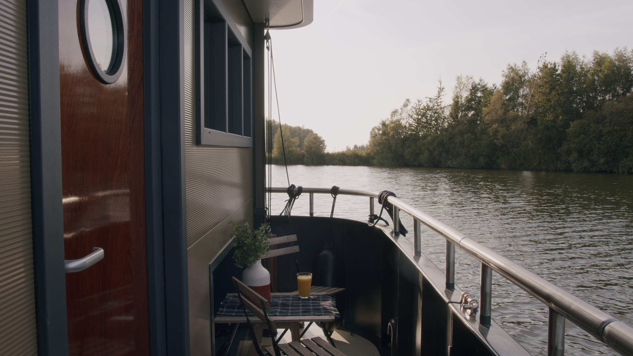 Boat deck with table and chairs, containing a glass of orange juice and a potted plant, overlooking a river and trees.