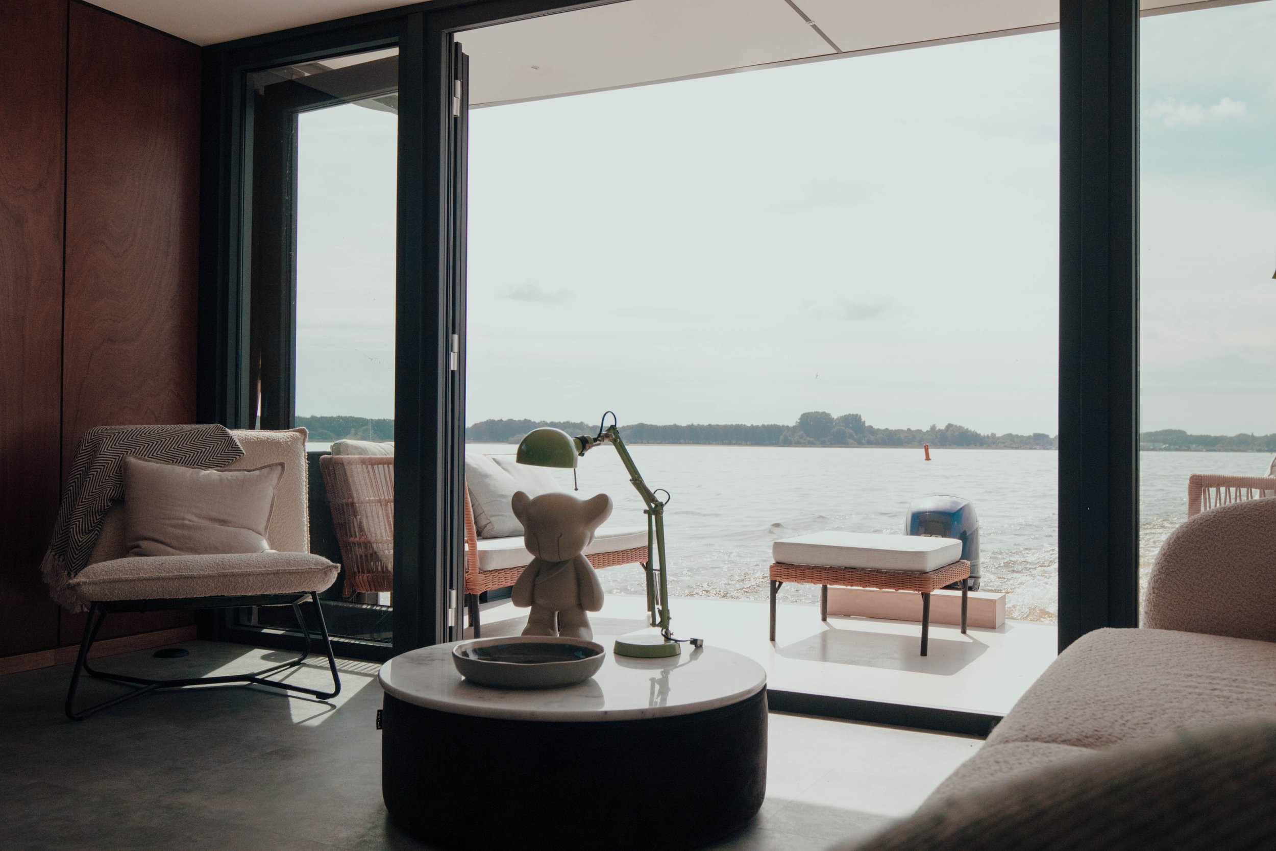 Modern living room with lake view, featuring armchairs, a small white sculpture, green desk lamp on a round table, and sliding glass doors.