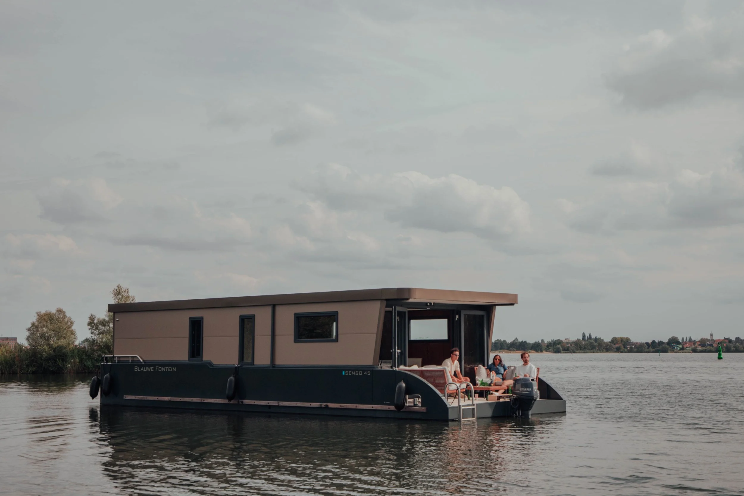 Modern houseboat with people lounging on the deck, floating on a calm lake with a scenic background.